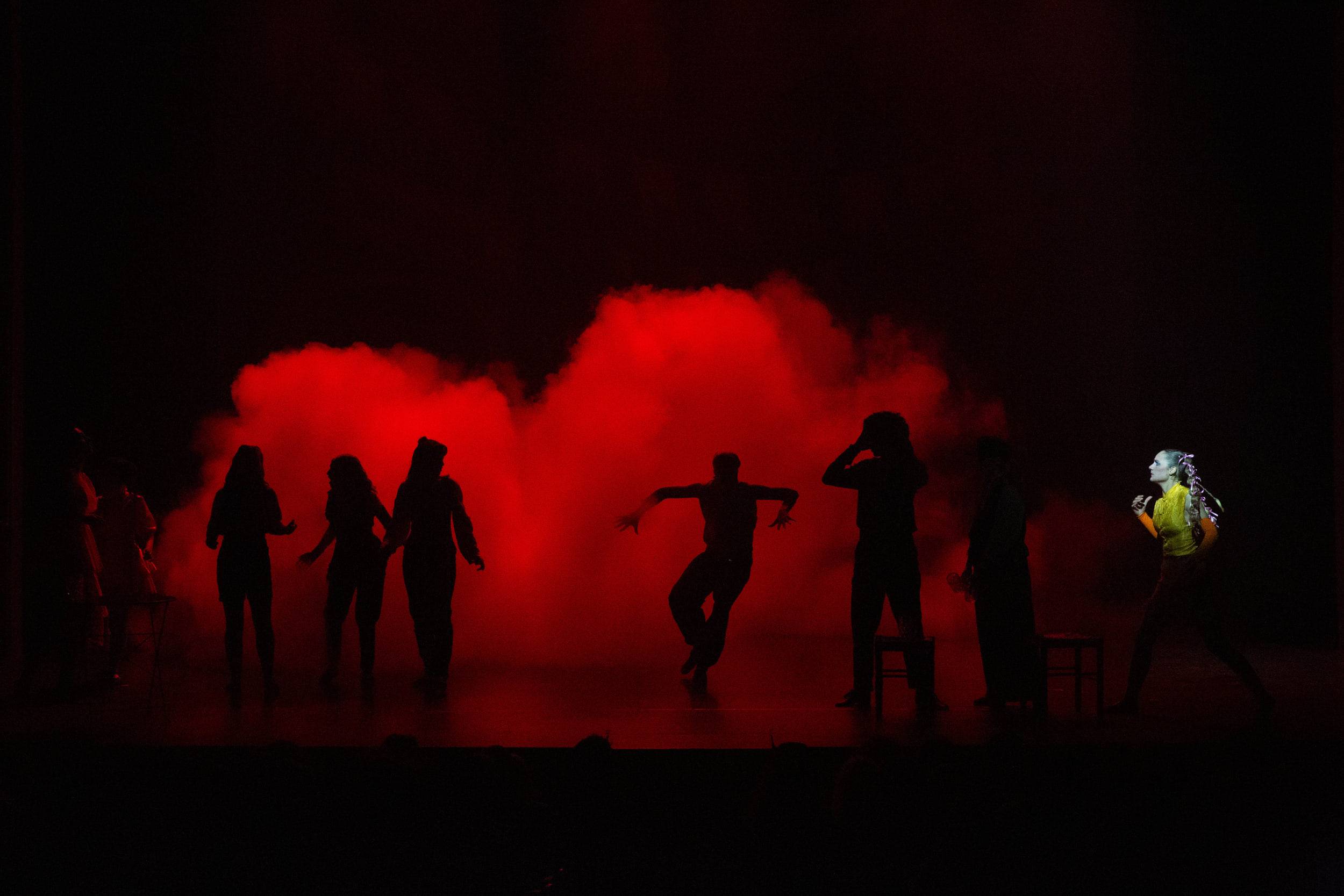 Des silhouettes de danseurs dans un nuage de fumée rouge, une seule figure éclairée à droite du cadre.