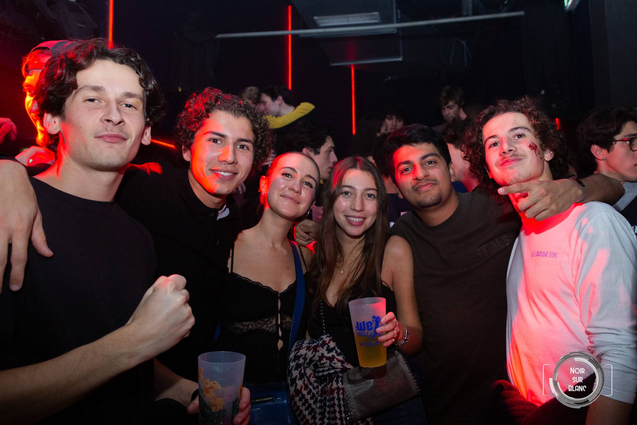 Un groupe serré face à l'objectif lève ses gobelets dans la lumière rose et rouge du bar.