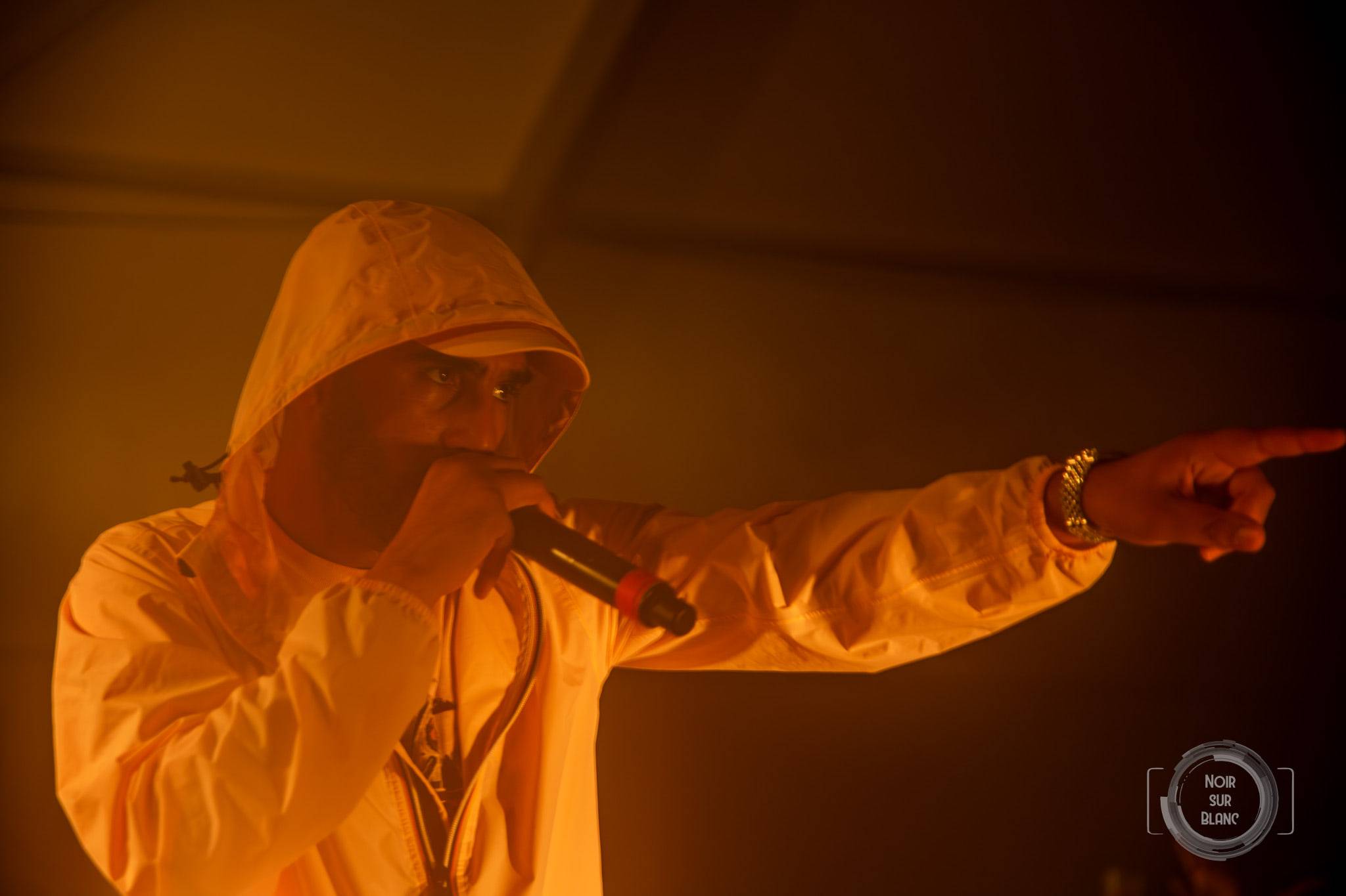 Baigné d'une lumière orange saturée, un artiste capuche relevée chante dans son micro en pointant le public du doigt.