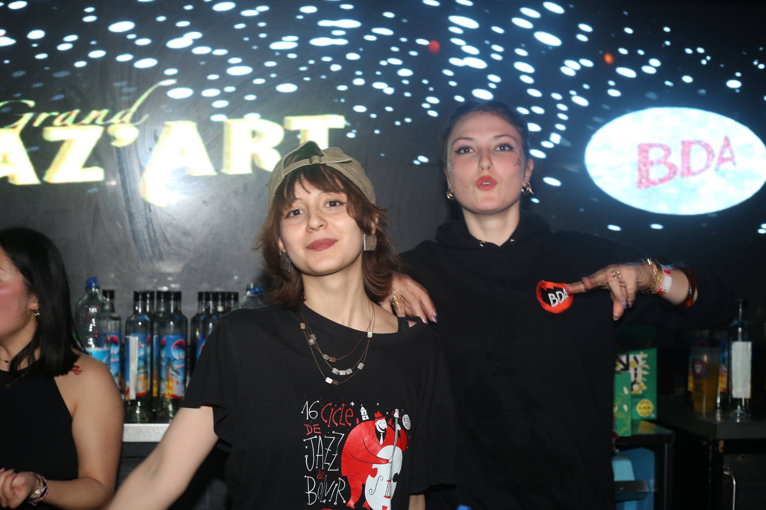 Deux étudiantes posent derrière le bar, sous les enseignes lumineuses « Grand Jaz'Art » et « BDA ».