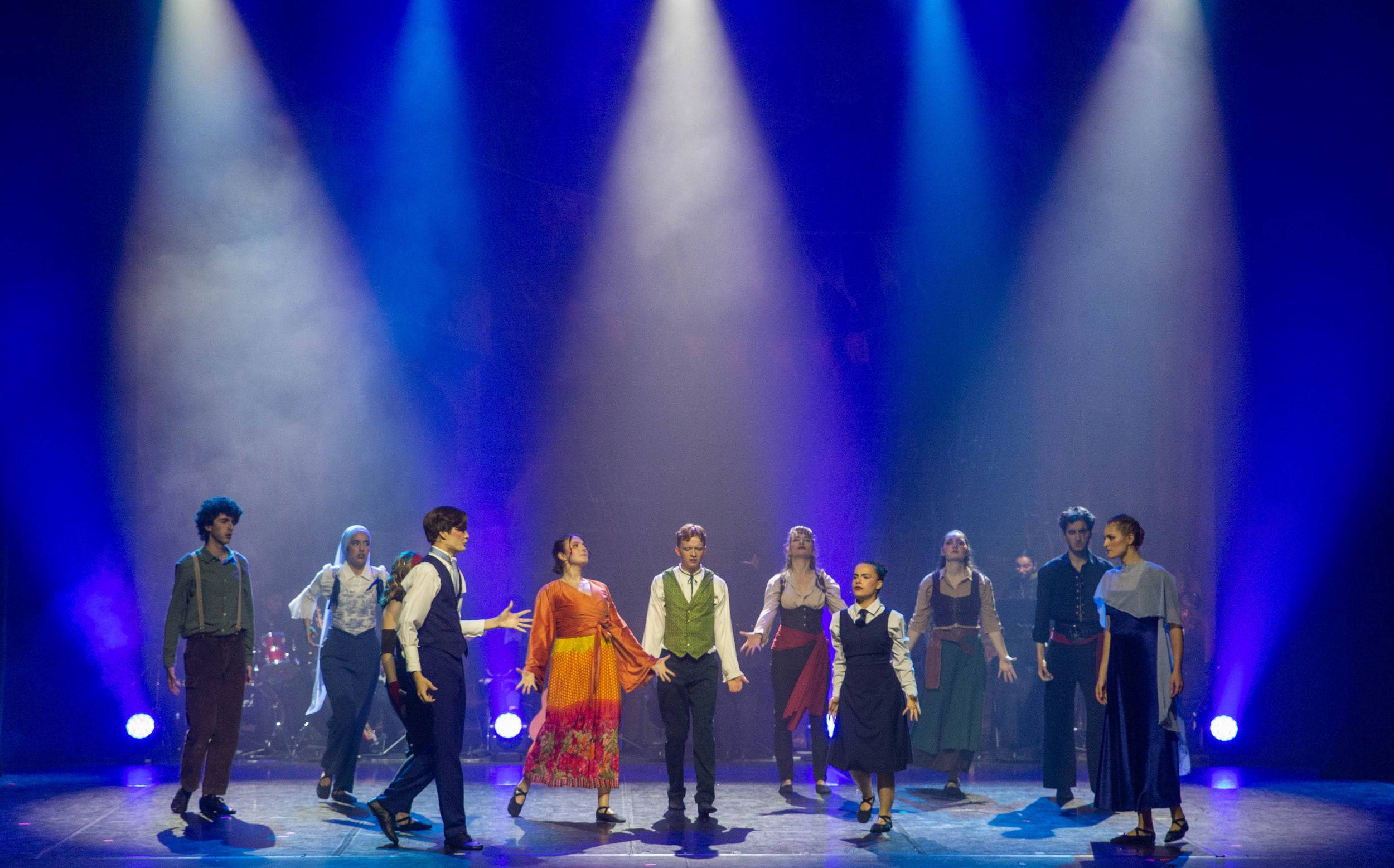 La troupe entière alignée face au public sous une rampe de projecteurs bleus.