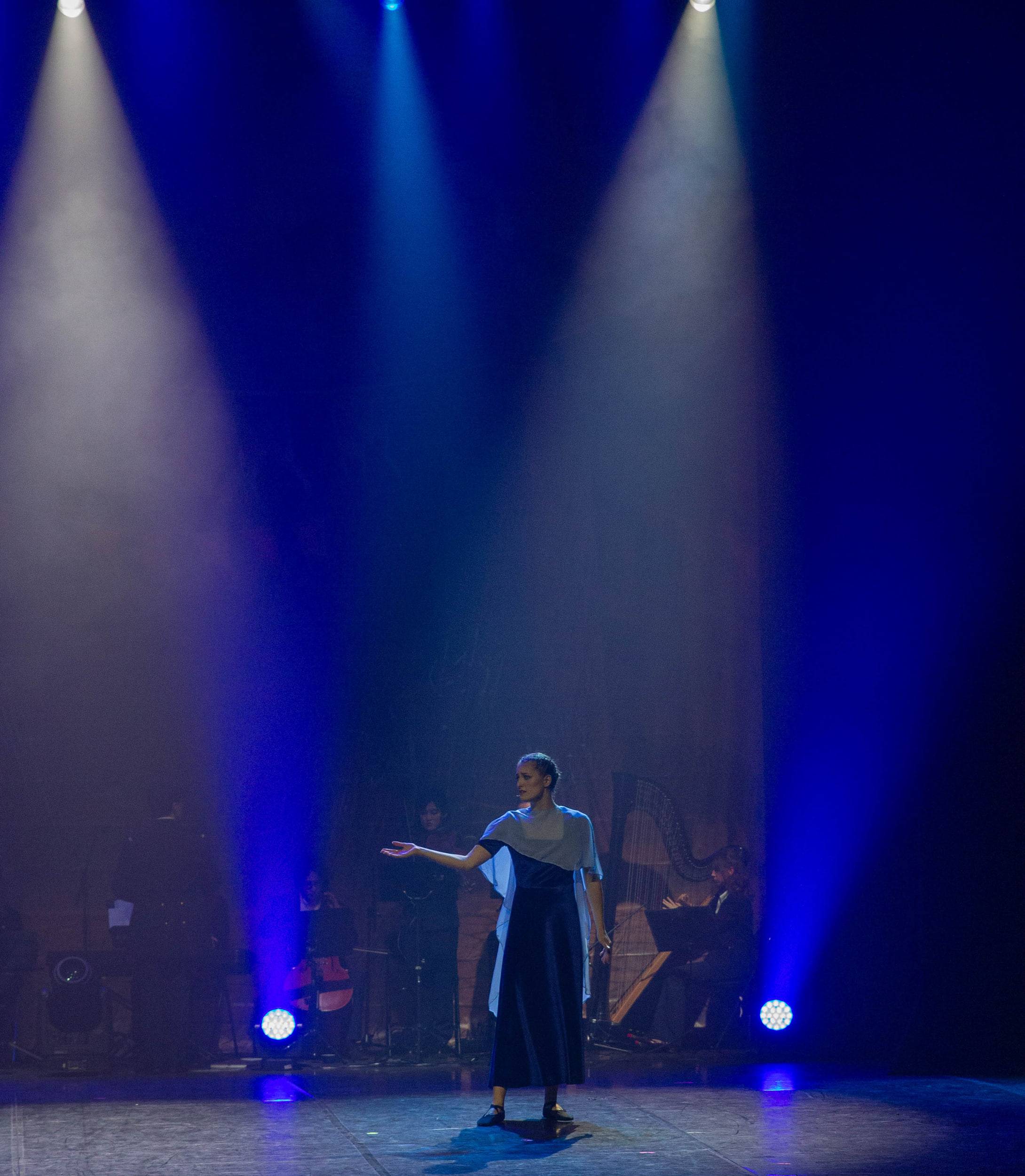Une comédienne seule au centre du plateau, un bras ouvert, l'orchestre en ombre derrière elle.
