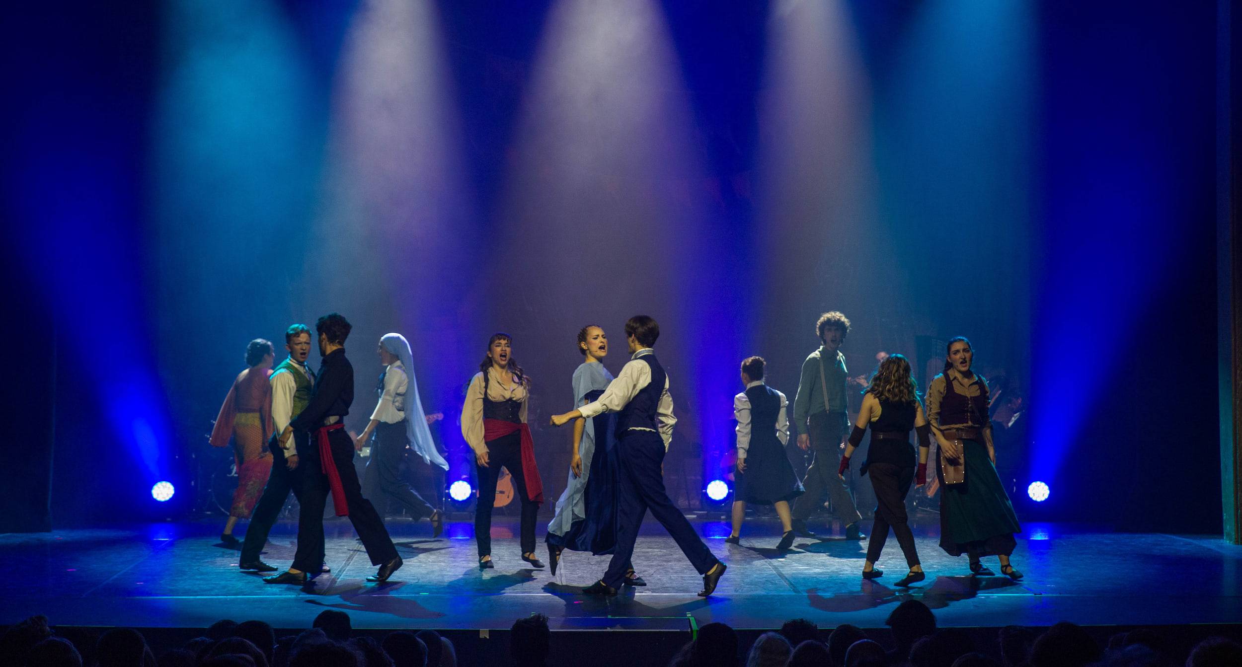 La troupe traverse le plateau en marchant, éclairée par une rangée de faisceaux bleus.