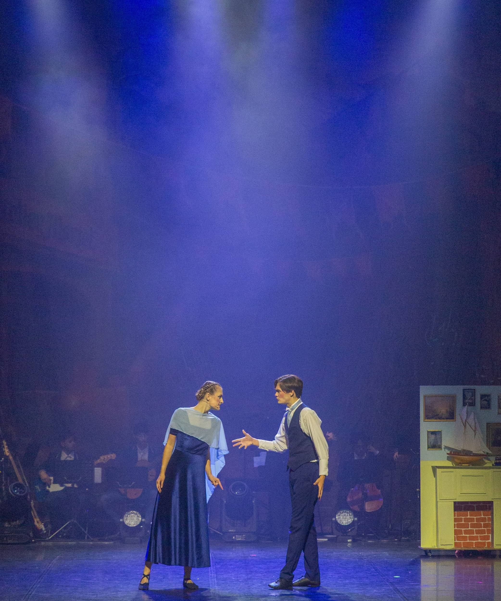 Un comédien tend la main vers une comédienne en robe bleue, au centre du plateau.
