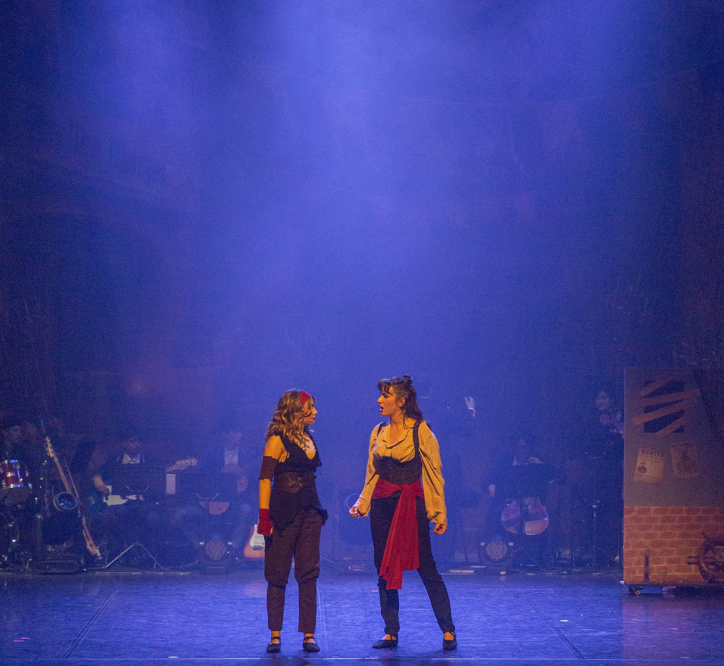 Deux comédiennes se font face au centre du plateau, l'orchestre jouant derrière elles.