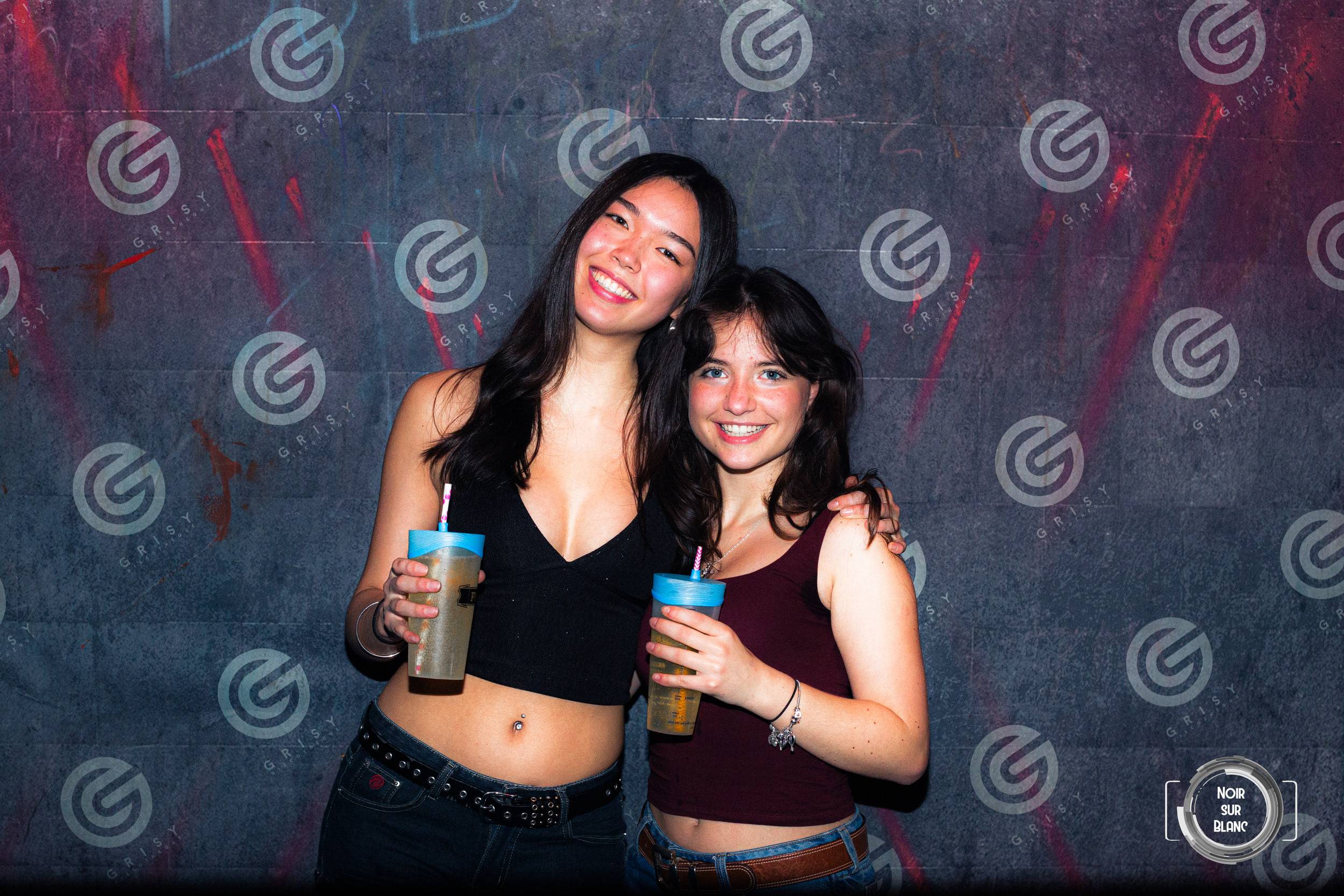 Deux étudiantes posent devant le mur logotypé de la discothèque, gobelets à la paille à la main.