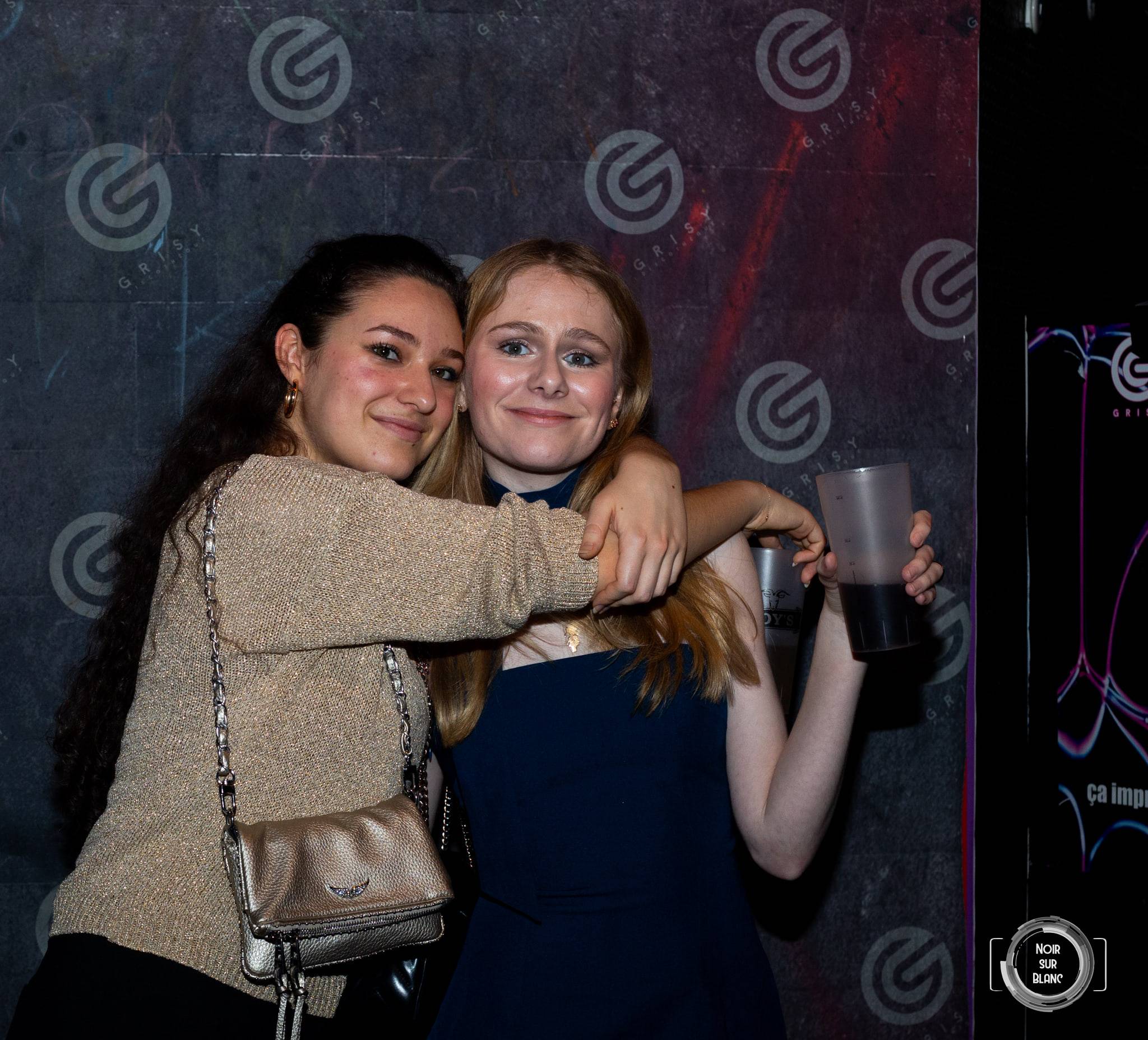 Deux amies s'enlacent en riant devant le mur logotypé de la discothèque, un gobelet noir à la main.
