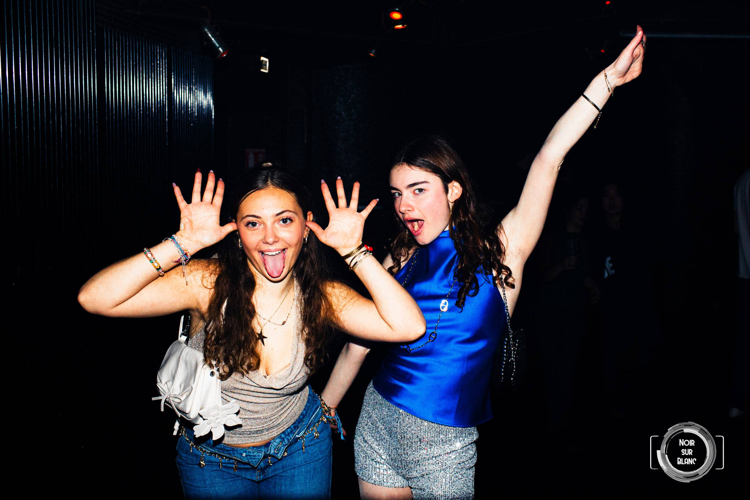 Deux étudiantes lèvent les bras et tirent la langue sur la piste de danse, saisies par un flash rasant.