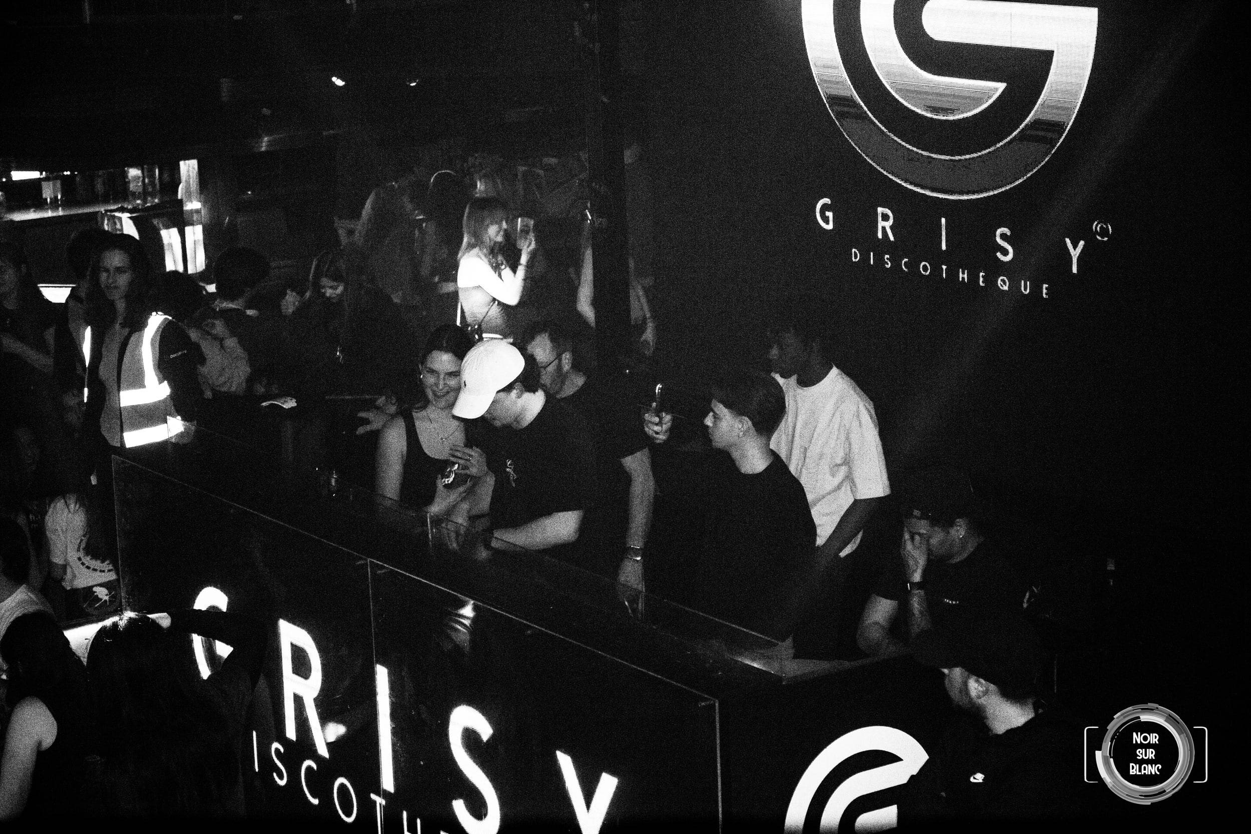 Vue en noir et blanc du long comptoir de la discothèque, la foule massée derrière, sous l'enseigne du club.