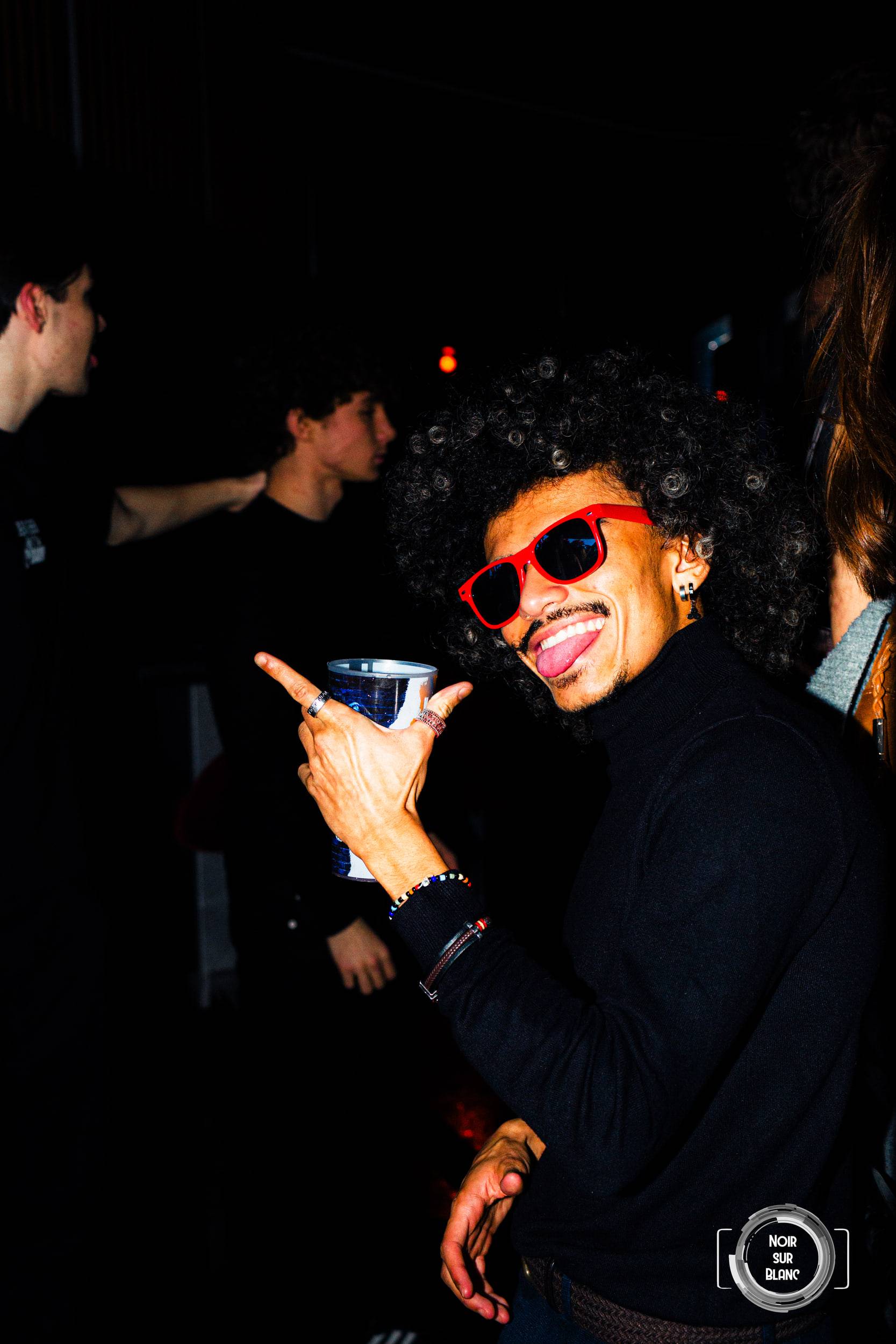 Un fêtard en col roulé noir, lunettes de soleil oranges et coupe afro, pointe l'objectif du doigt, gobelet à la main.
