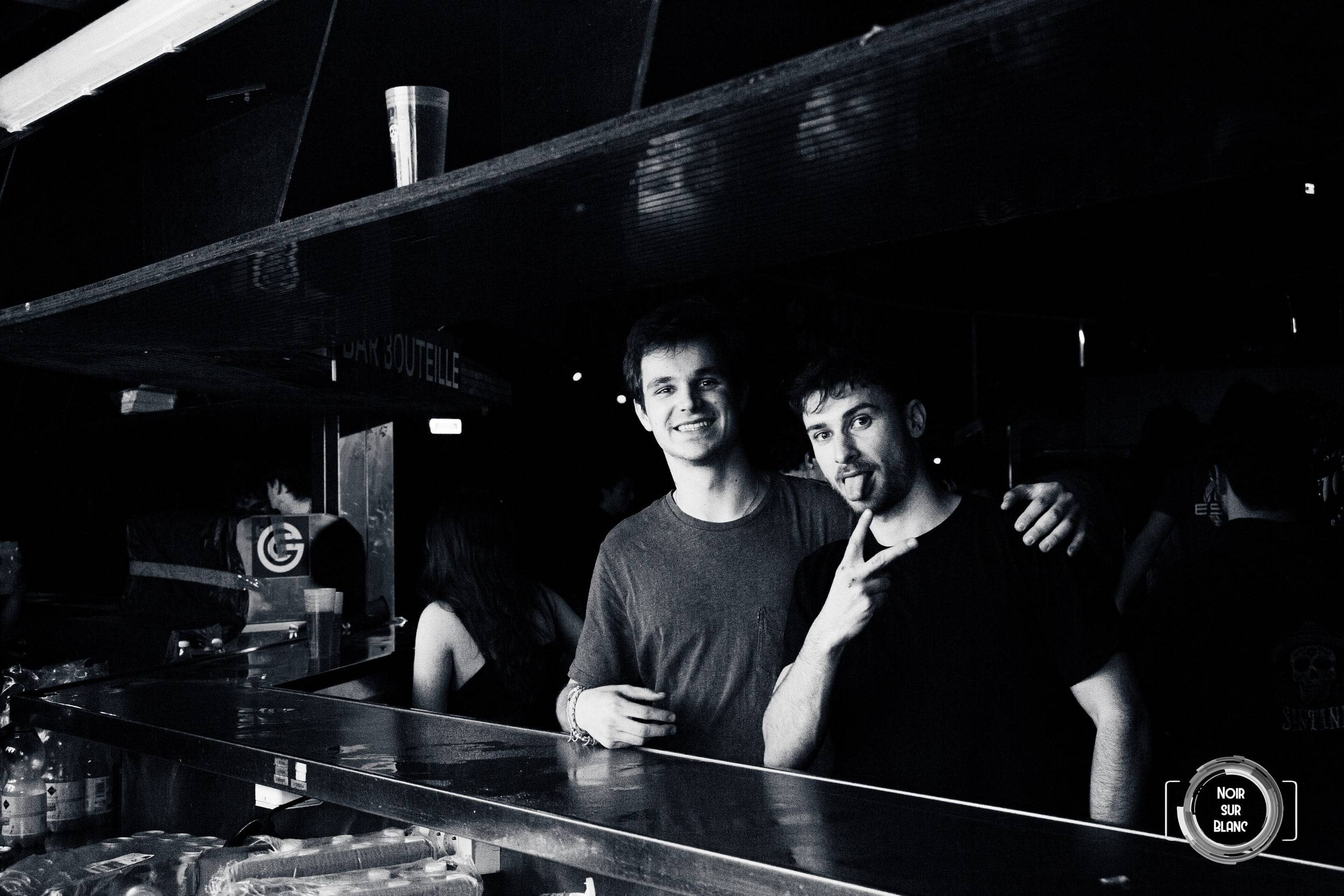 Photographie en noir et blanc de deux amis accoudés au comptoir du club, l'un faisant un signe de la main vers l'objectif.
