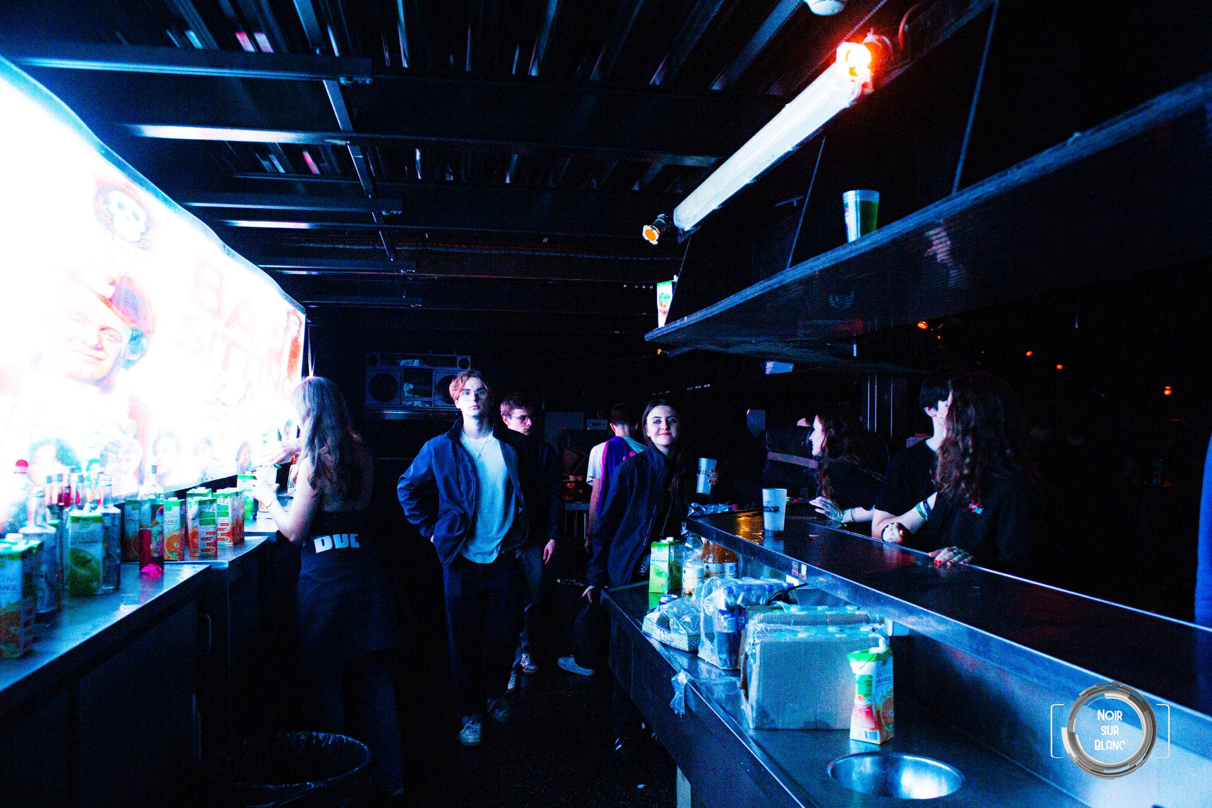 Le bar du club en enfilade sous une lumière bleue, bouteilles et bacs à glace alignés, quelques silhouettes au fond.