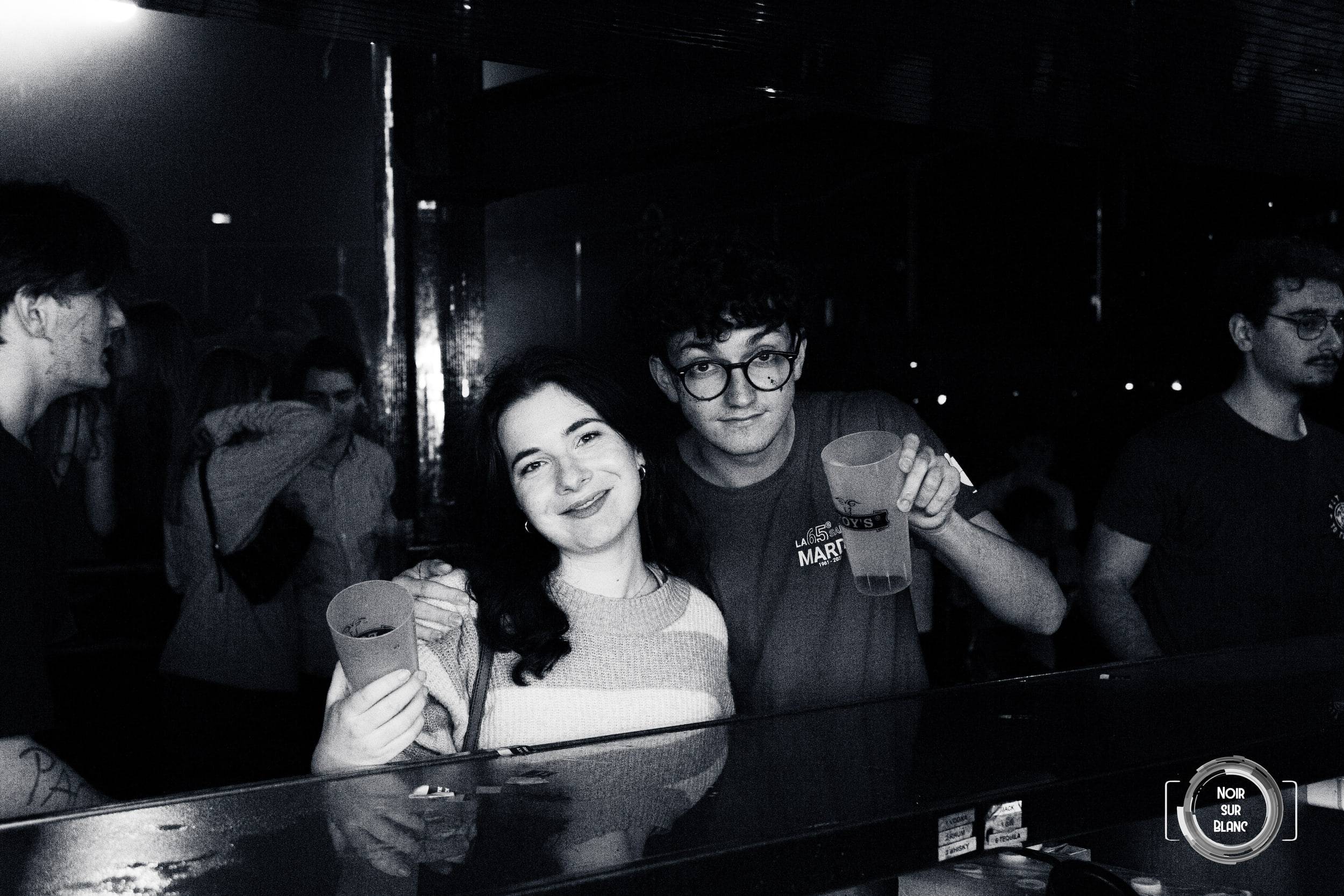 Photographie en noir et blanc de deux amis souriants derrière le comptoir de la discothèque, un gobelet à la main.