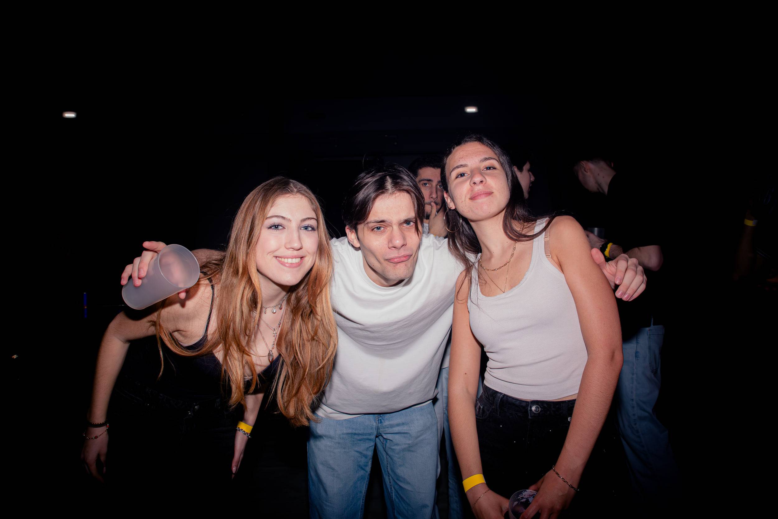 Trois étudiants serrés sourient face à l'objectif, un bracelet d'entrée jaune au poignet.