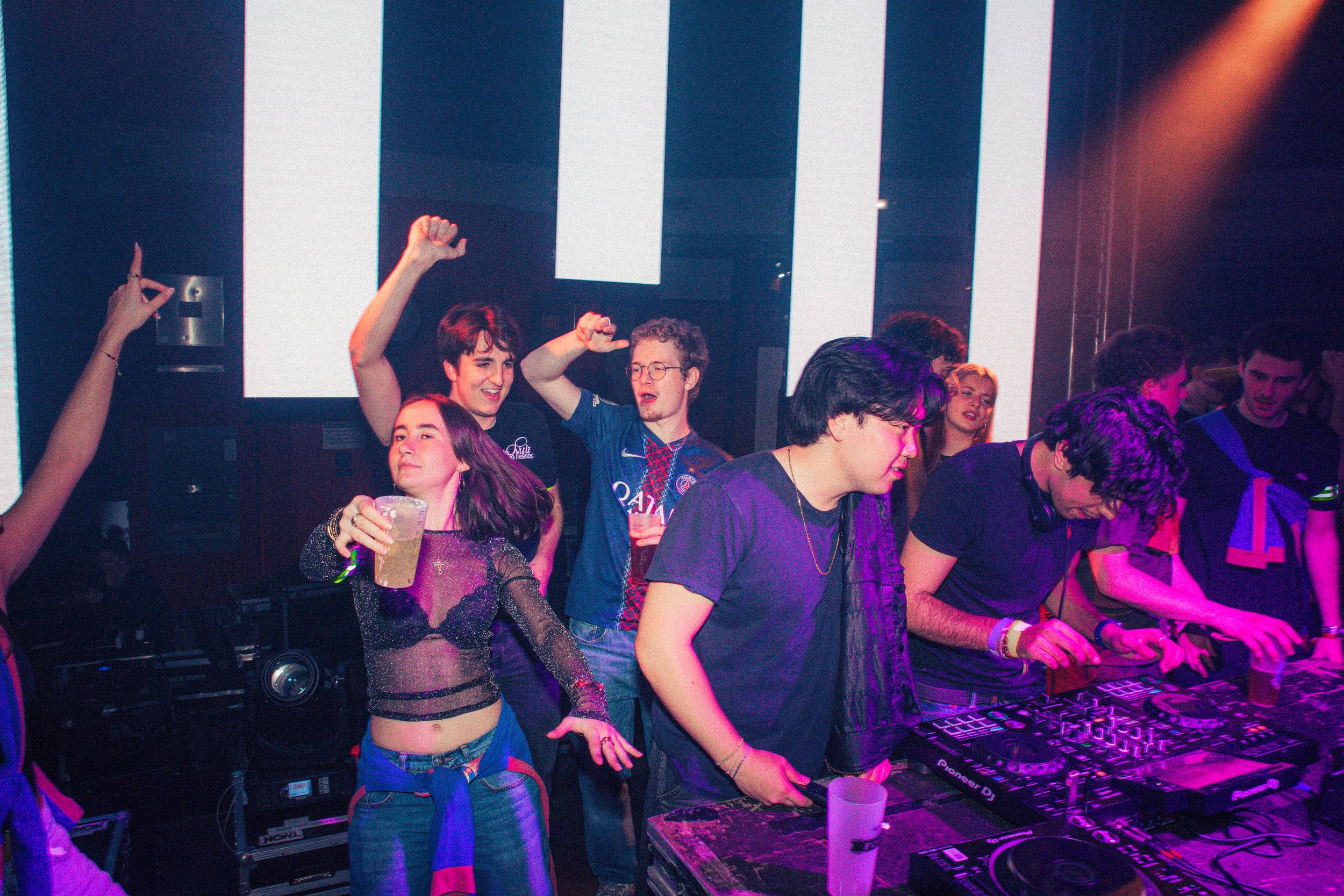 Un groupe danse derrière la console du DJ devant un mur d'écrans, dans une lumière violette.