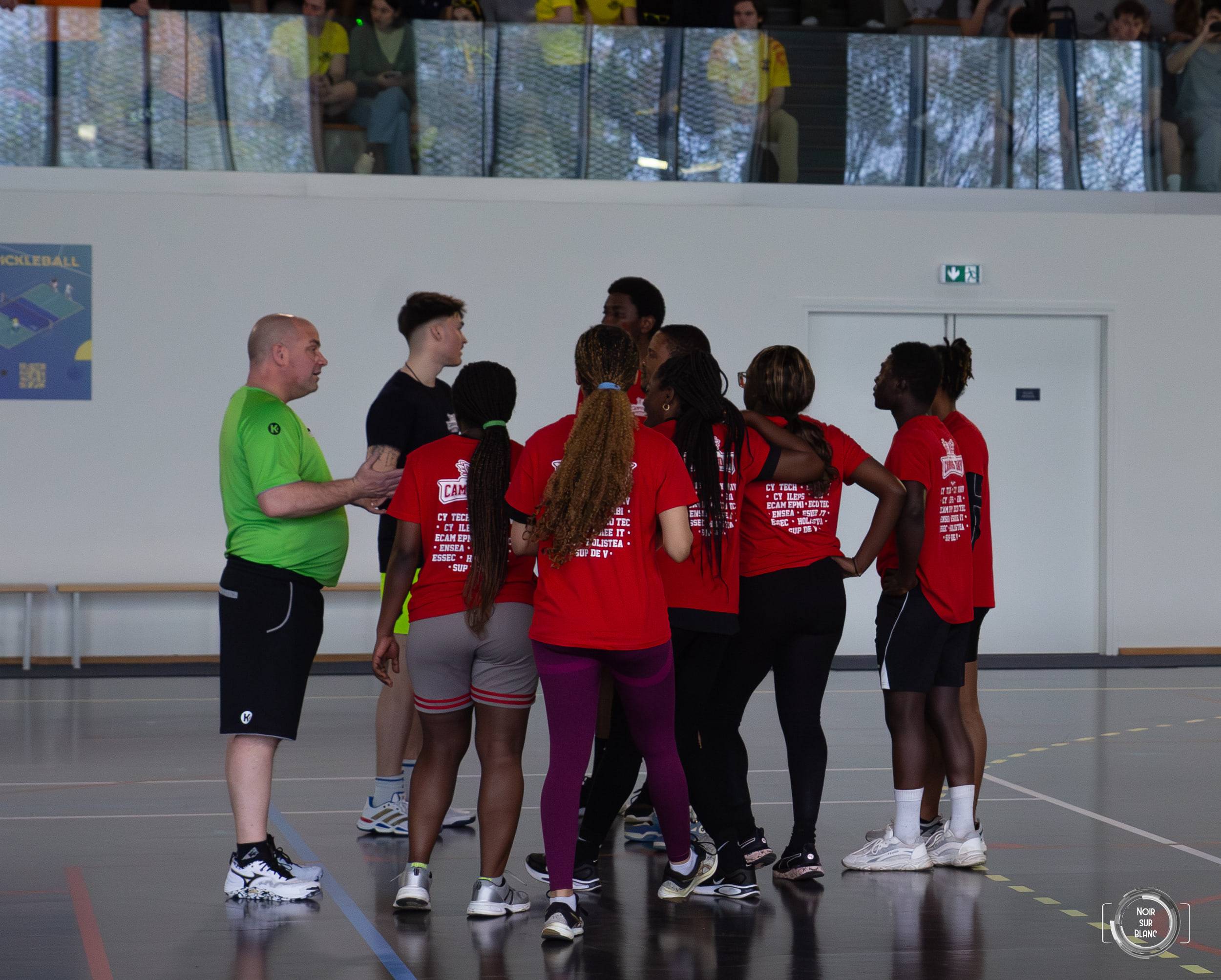 Un entraîneur en chasuble verte rassemble en cercle une équipe en t-shirts rouges avant le coup d'envoi.