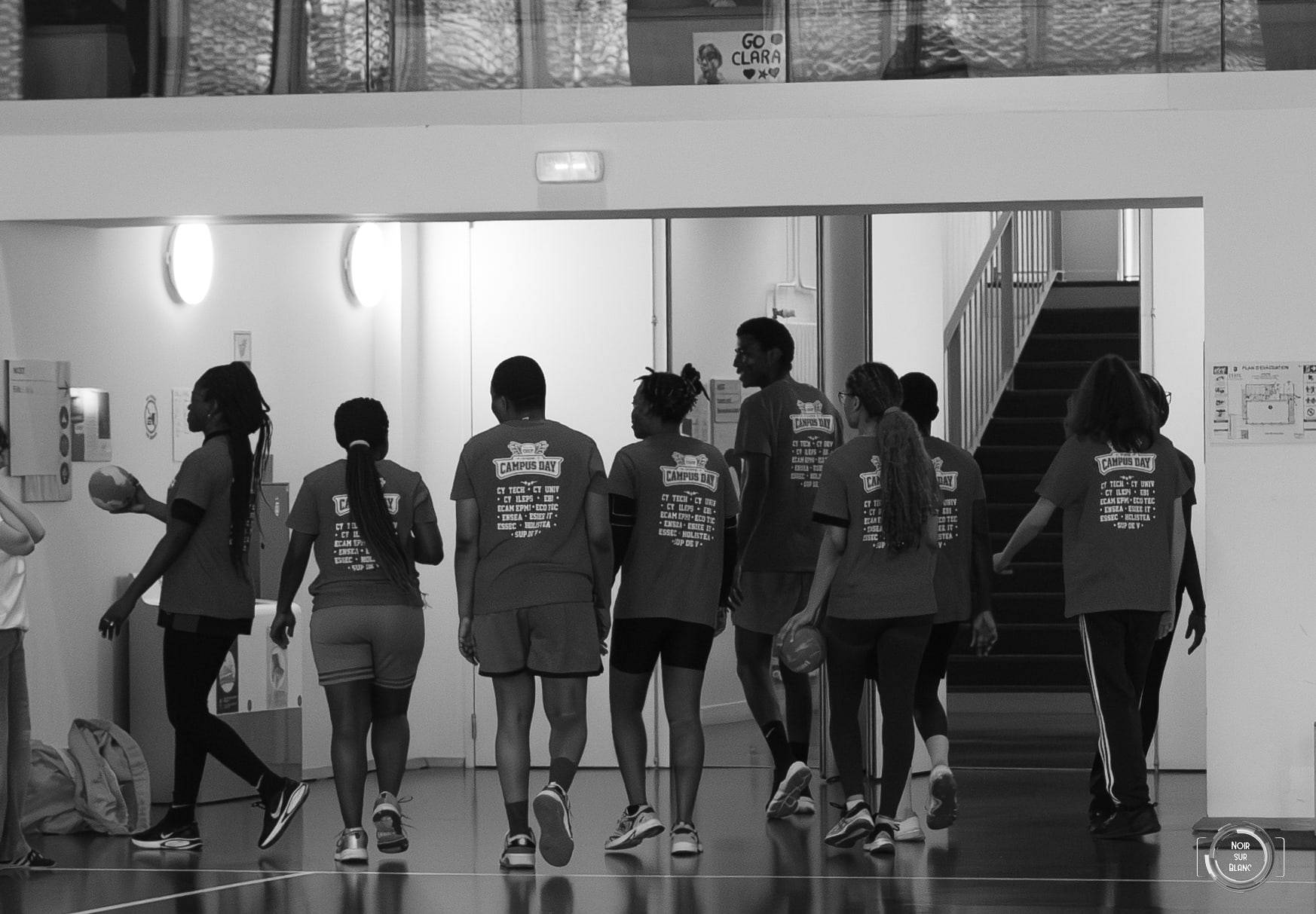 En noir et blanc, une équipe en t-shirts floqués marche de dos dans un couloir vitré, en route vers le gymnase.