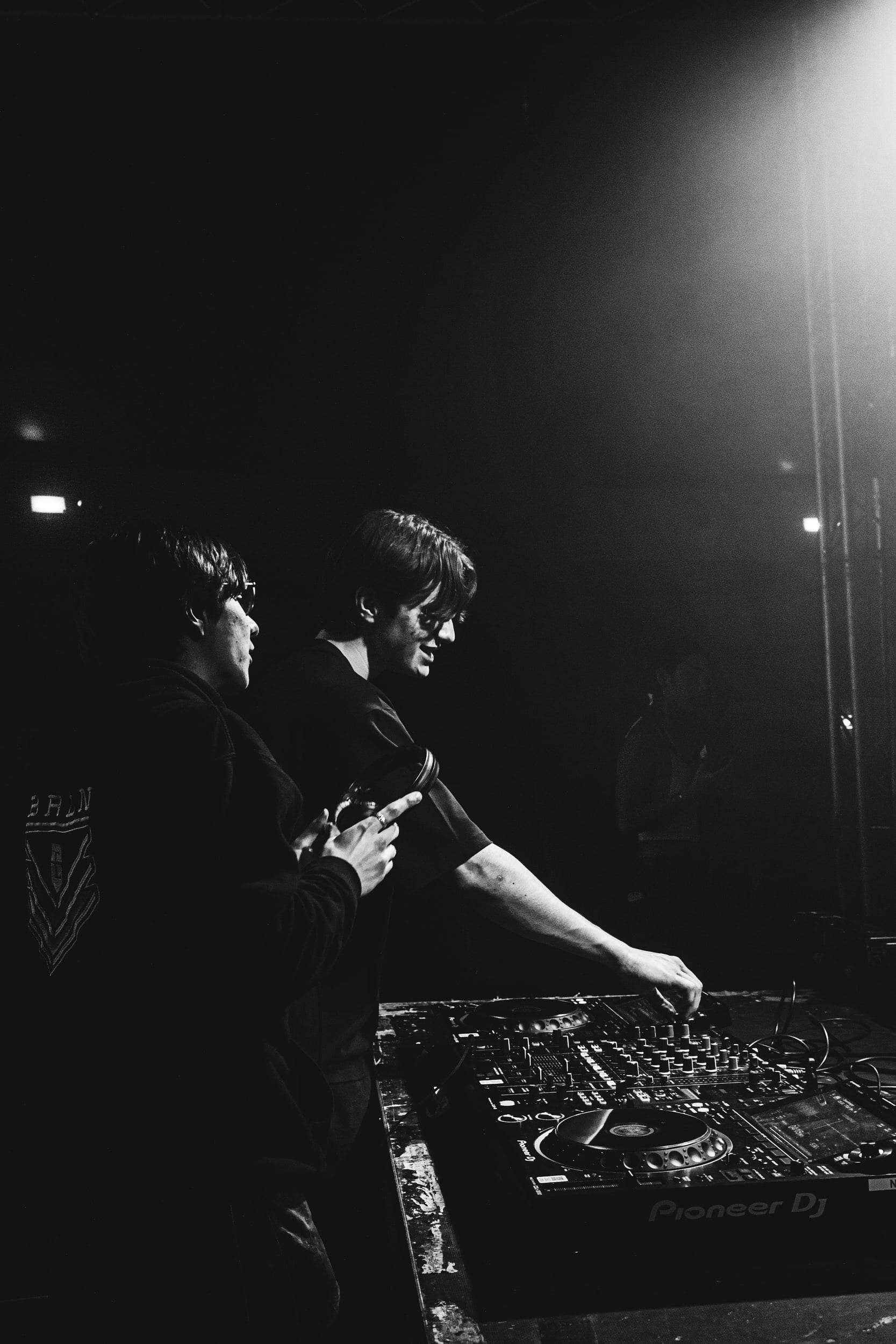 Photographie en noir et blanc de deux DJ penchés sur leur console, un faisceau de lumière au-dessus d'eux.