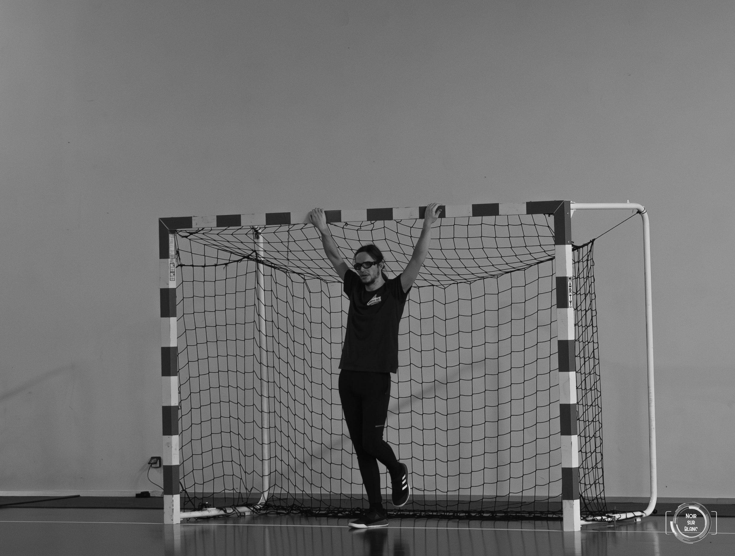 En noir et blanc, un gardien de handball attend seul dans sa cage, bras écartés, sur un fond parfaitement vide.