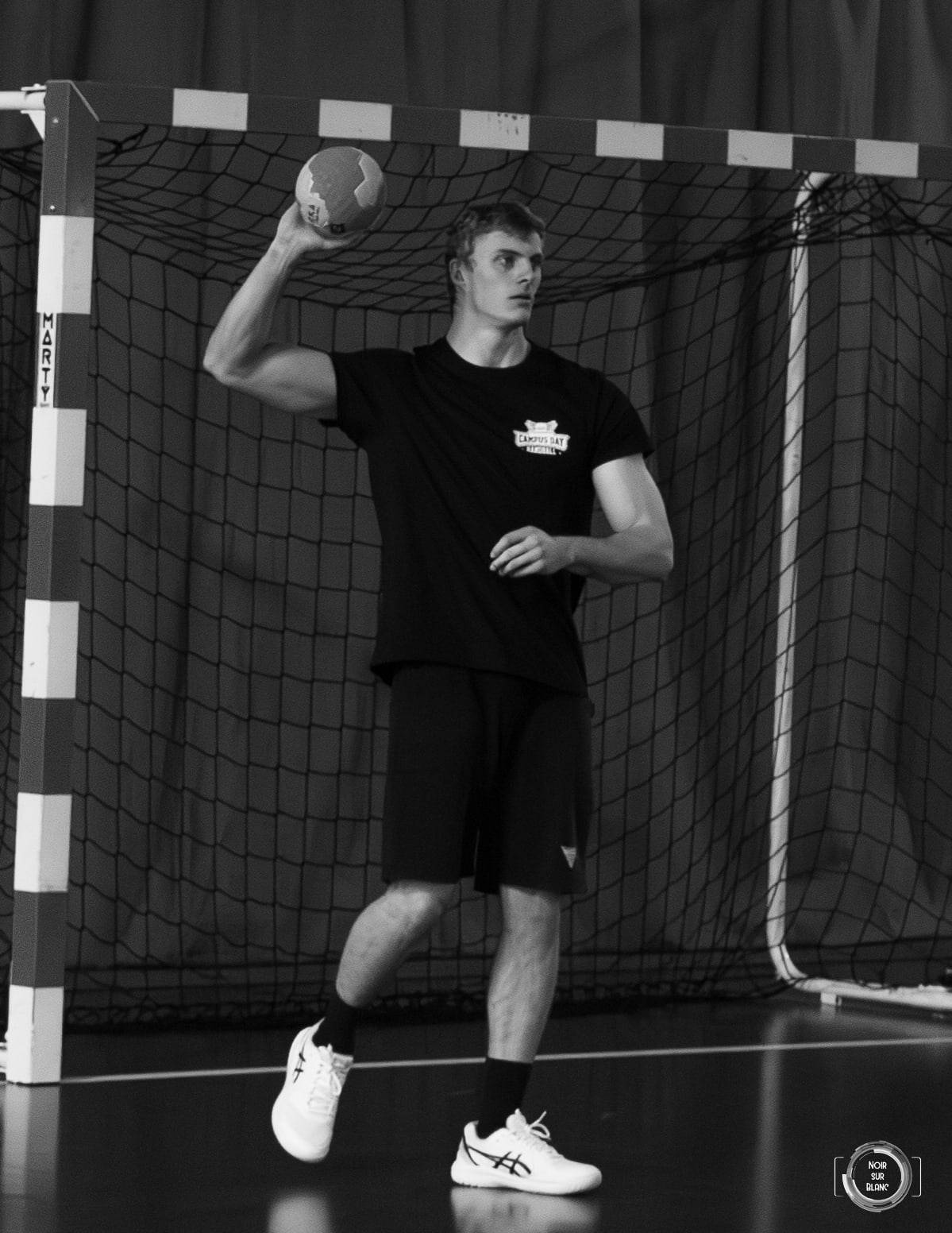 En noir et blanc, un handballeur arme son tir devant une cage vide, seul dans le gymnase.