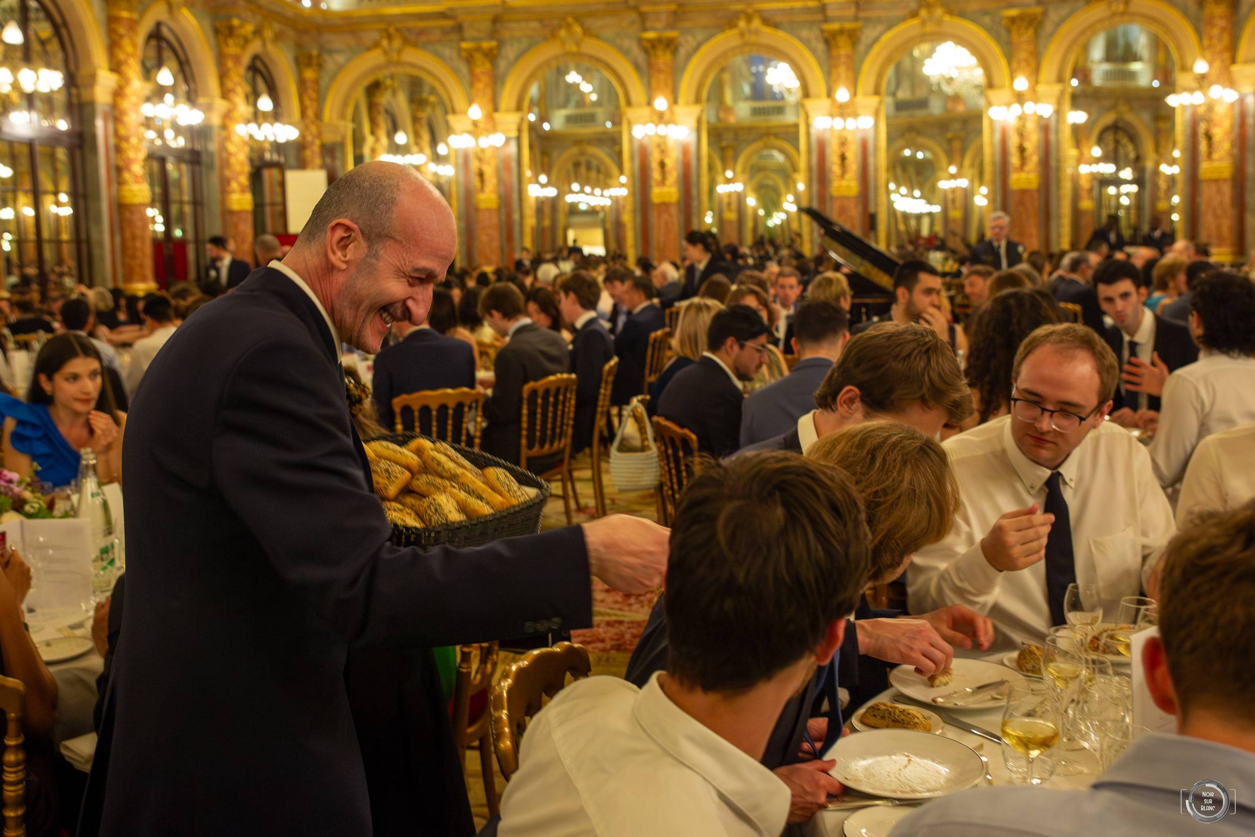 Vue d'ensemble d'une salle de bal dorée aux lustres de cristal, remplie de convives attablés en tenue de soirée.
