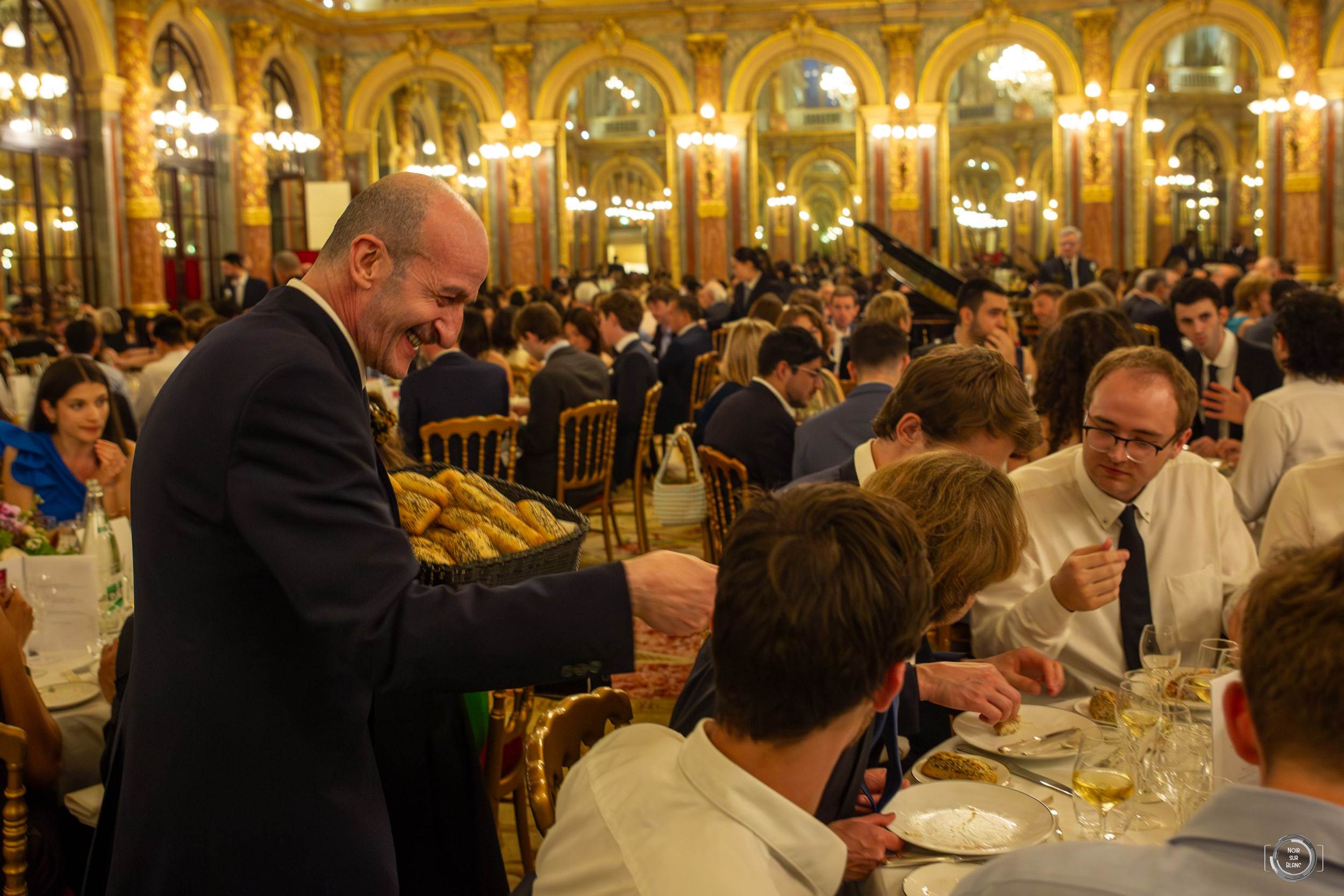 Vue d'ensemble d'une salle de bal dorée aux lustres de cristal, remplie de convives attablés en tenue de soirée.