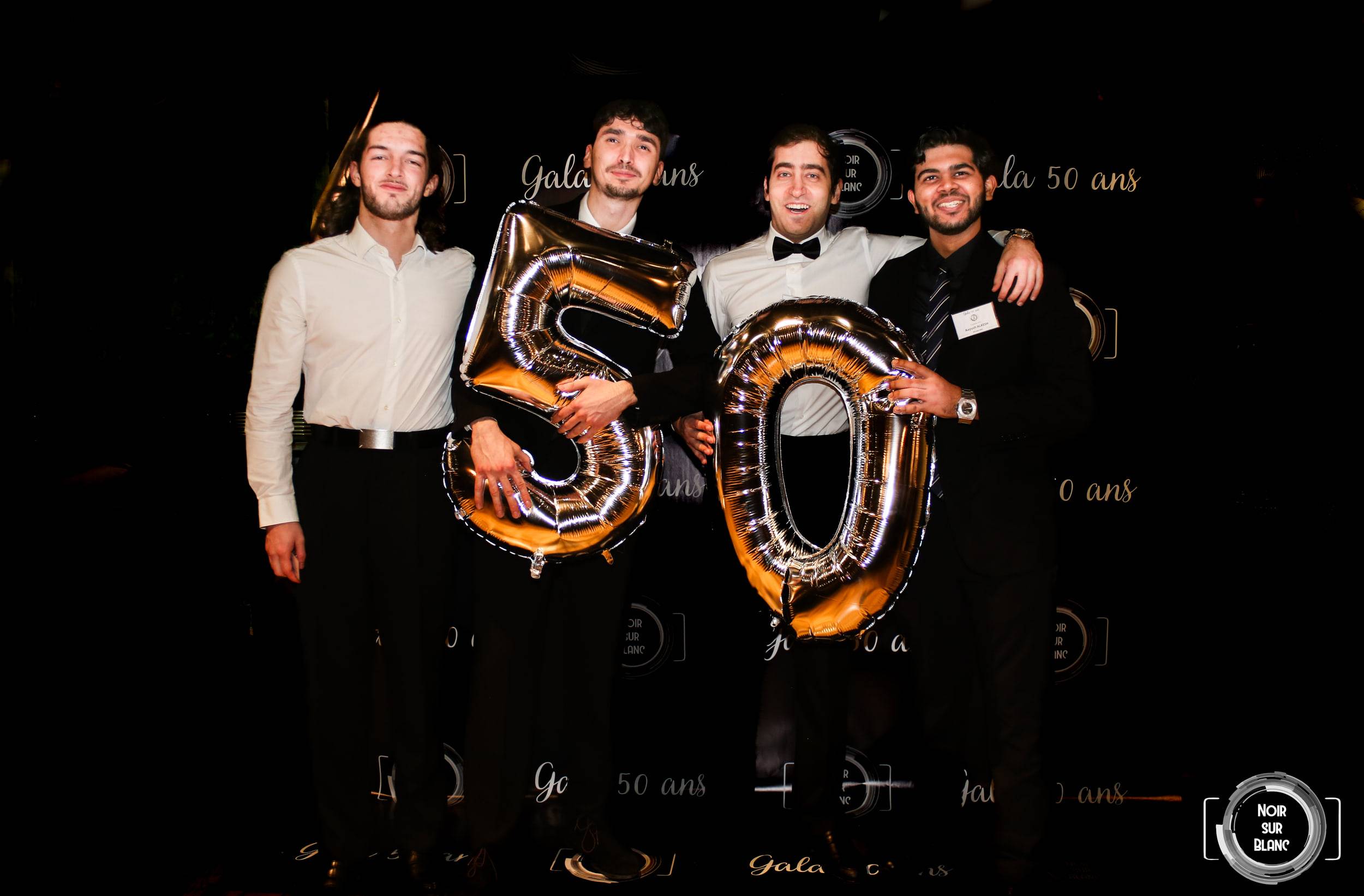 Quatre invités en tenue de soirée brandissent deux ballons dorés en forme de 5 et de 0 devant une bâche répétant « Gala 50 ans ».
