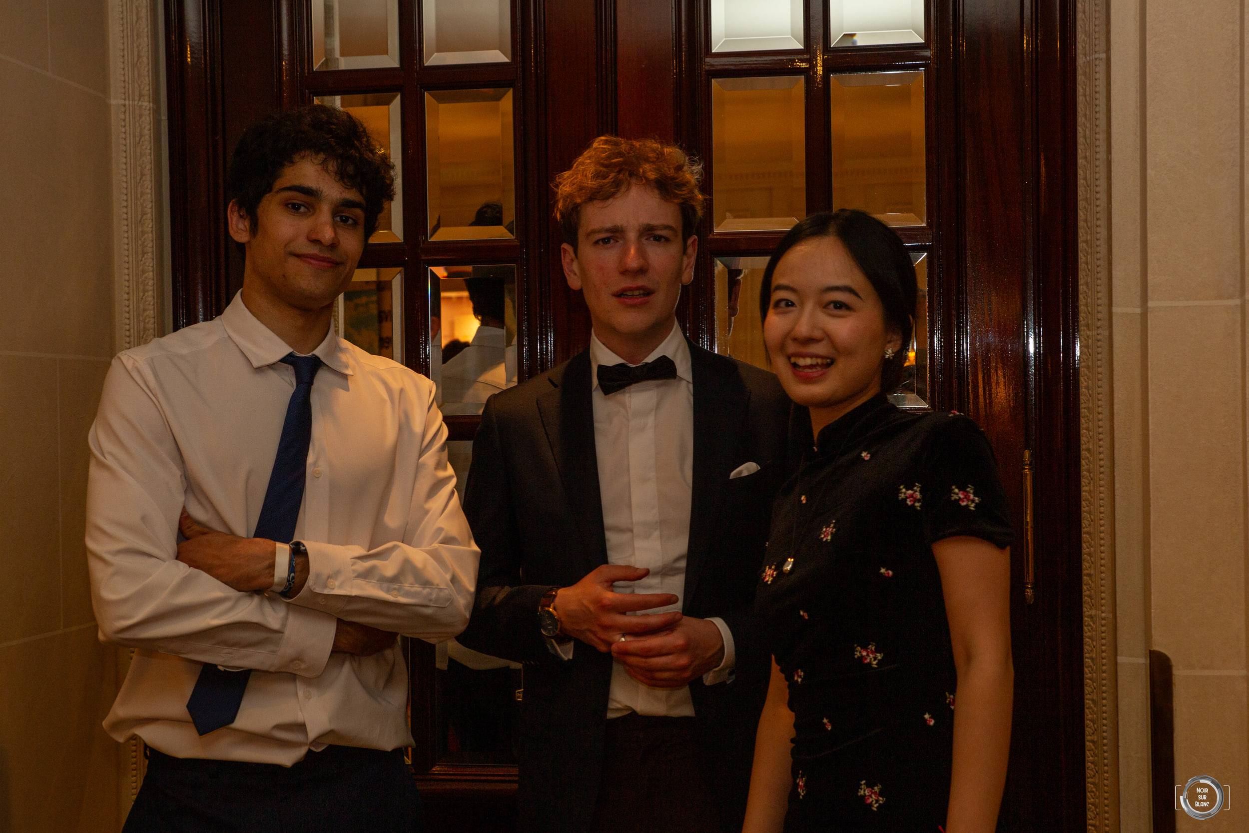 Trois étudiants en tenue de soirée, verre à la main, posent devant les boiseries et les portes vitrées d'un salon.