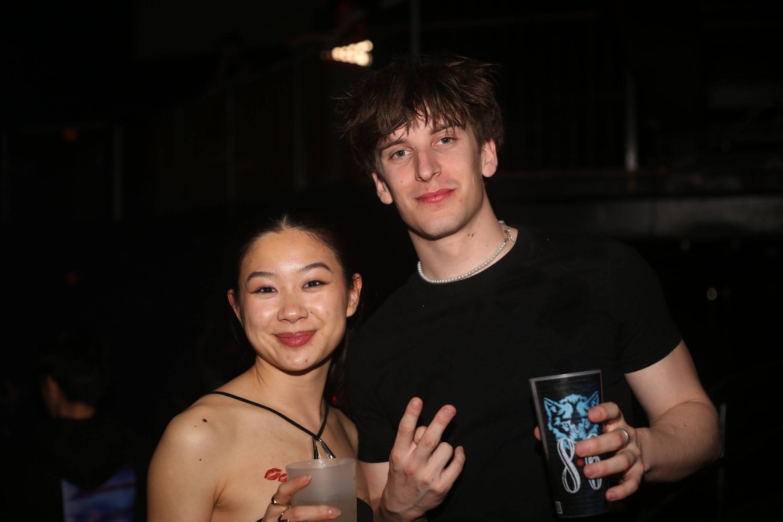 Deux amis posent dans la pénombre du club, l'un tient un gobelet, l'autre fait le signe de la victoire.