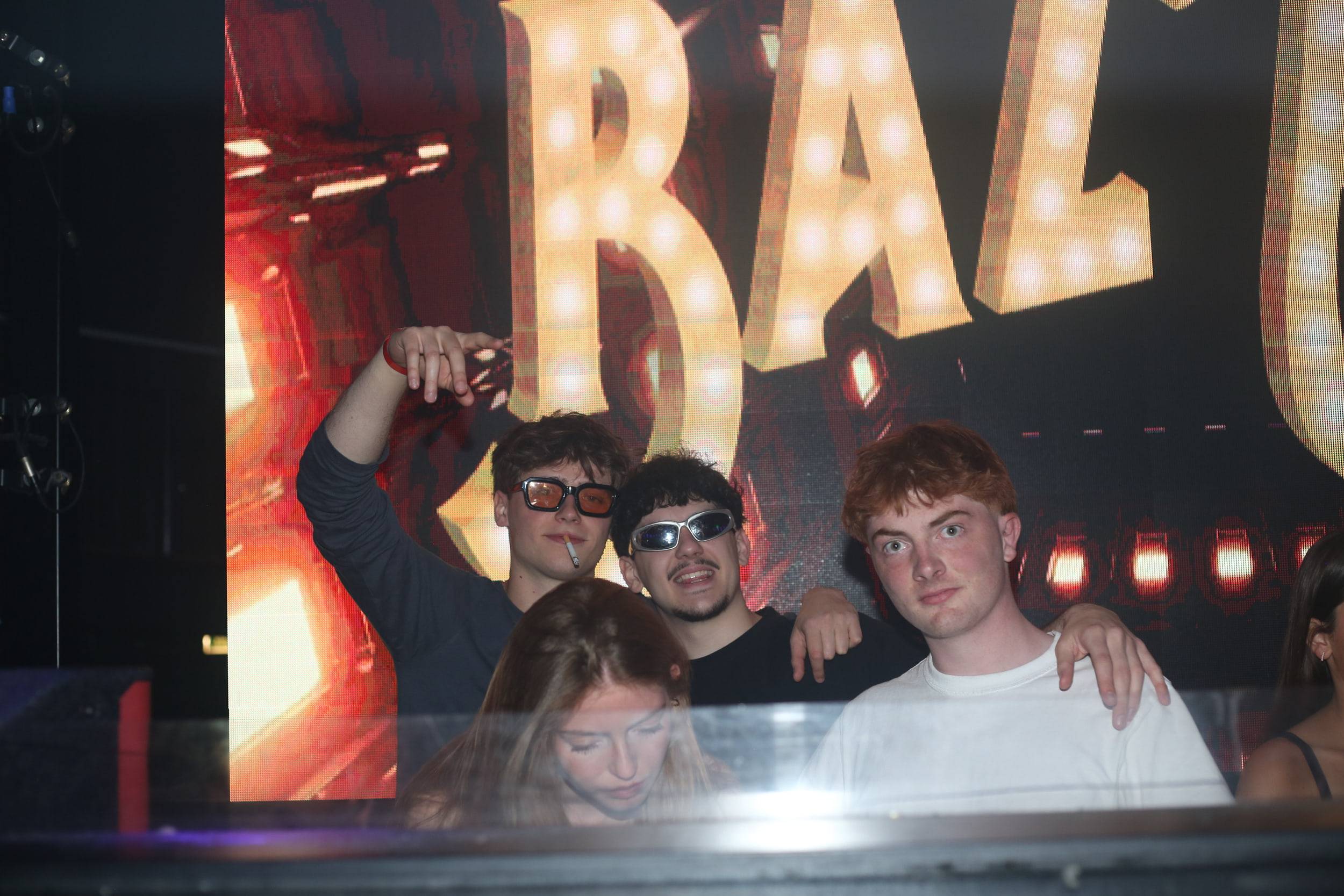 Trois fêtards posent devant une enseigne lumineuse à ampoules, deux d'entre eux en lunettes de soleil.