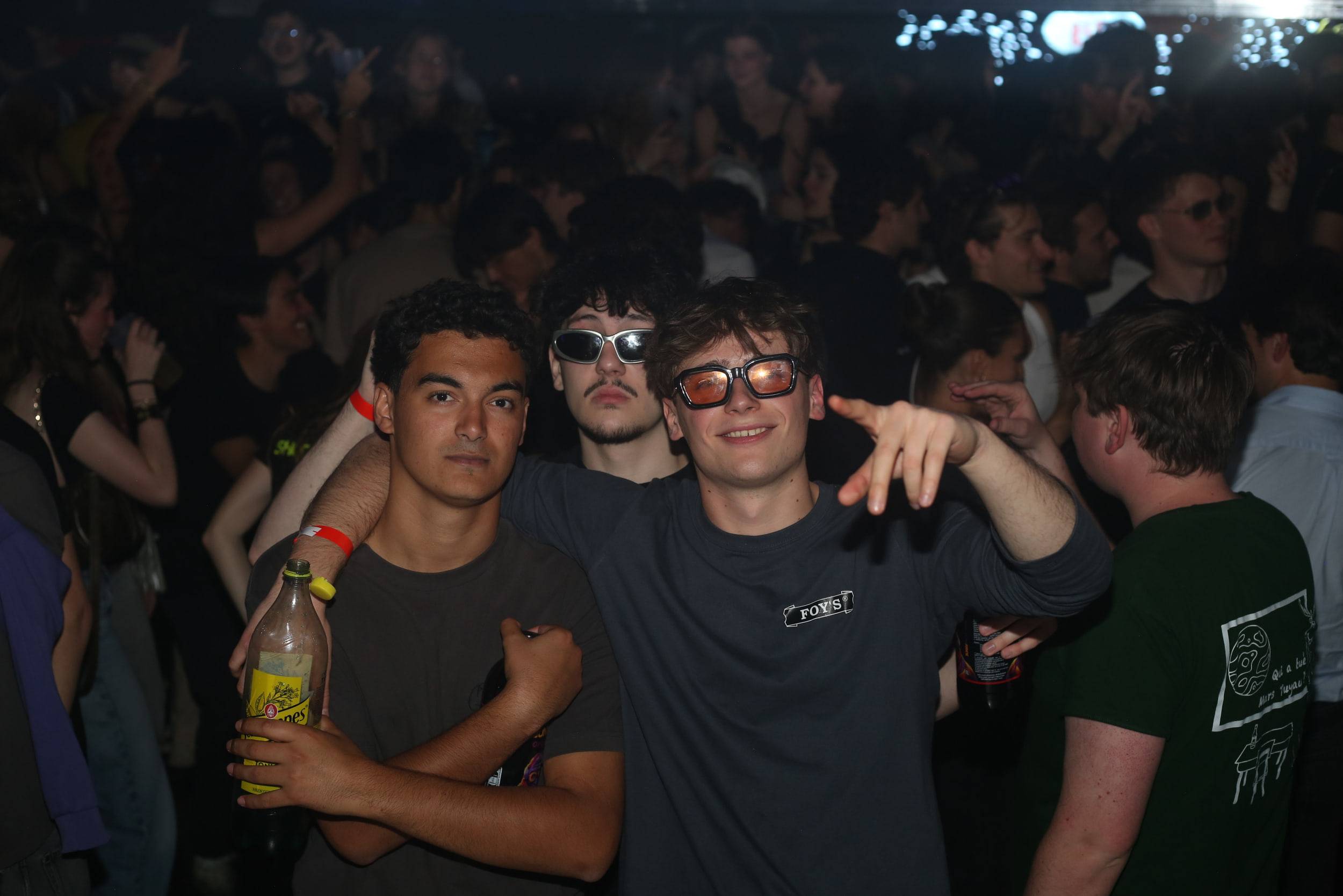 Trois étudiants posent en pleine foule, l'un en lunettes de soleil, une bouteille à la main.