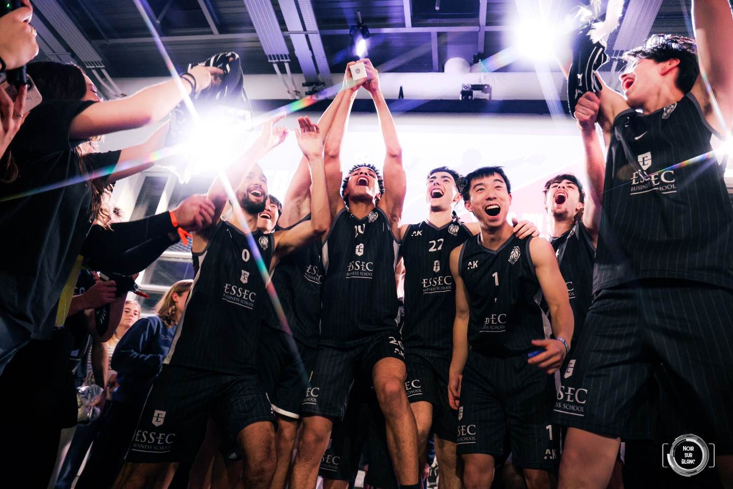 Les joueurs de l'équipe de basket de l'ESSEC lèvent un trophée en criant leur joie, éclairés par un flash en contre-jour.