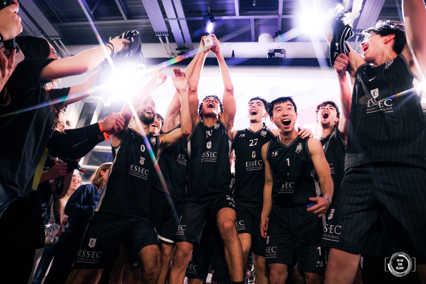 Les joueurs de l'équipe de basket de l'ESSEC lèvent un trophée en criant leur joie, éclairés par un flash en contre-jour.