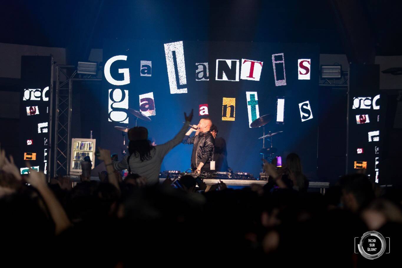 Un artiste sur scène, bras levé et micro à la main, devant un décor de lettres découpées épelant Galantis, au-dessus d'une foule.