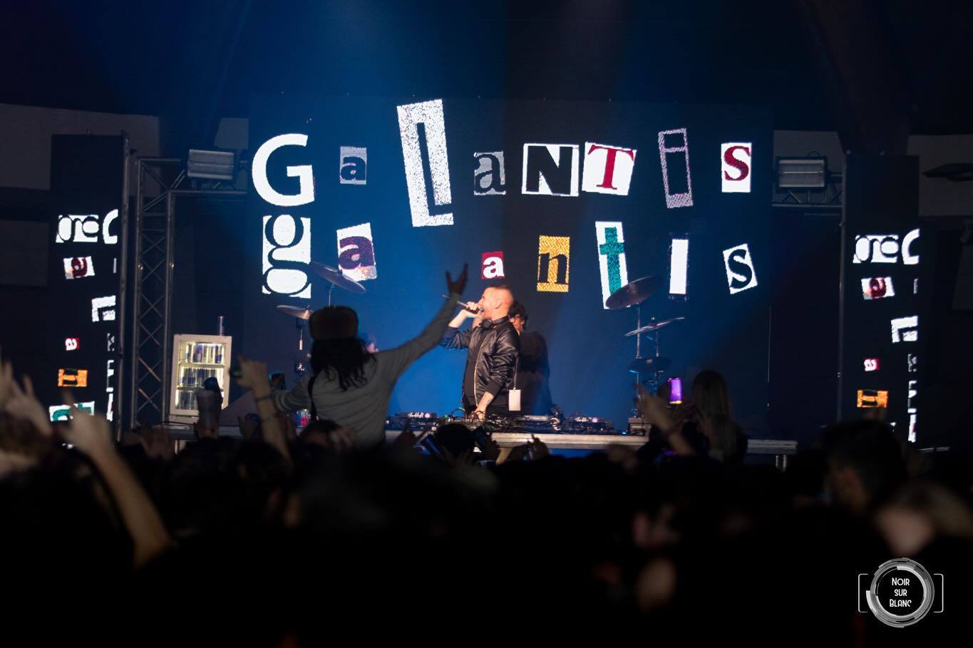 Un artiste sur scène, bras levé et micro à la main, devant un décor de lettres découpées épelant Galantis, au-dessus d'une foule.