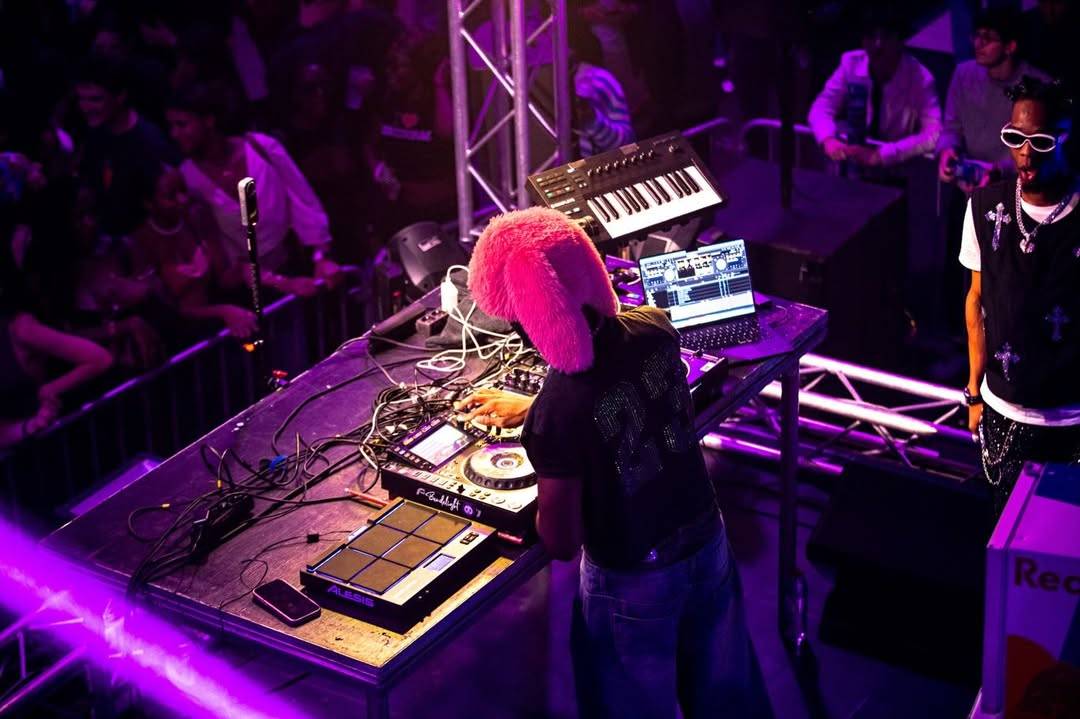 Vue en plongée d'un DJ coiffé d'une toque rose, penché sur ses platines sous une lumière violette, face au public.