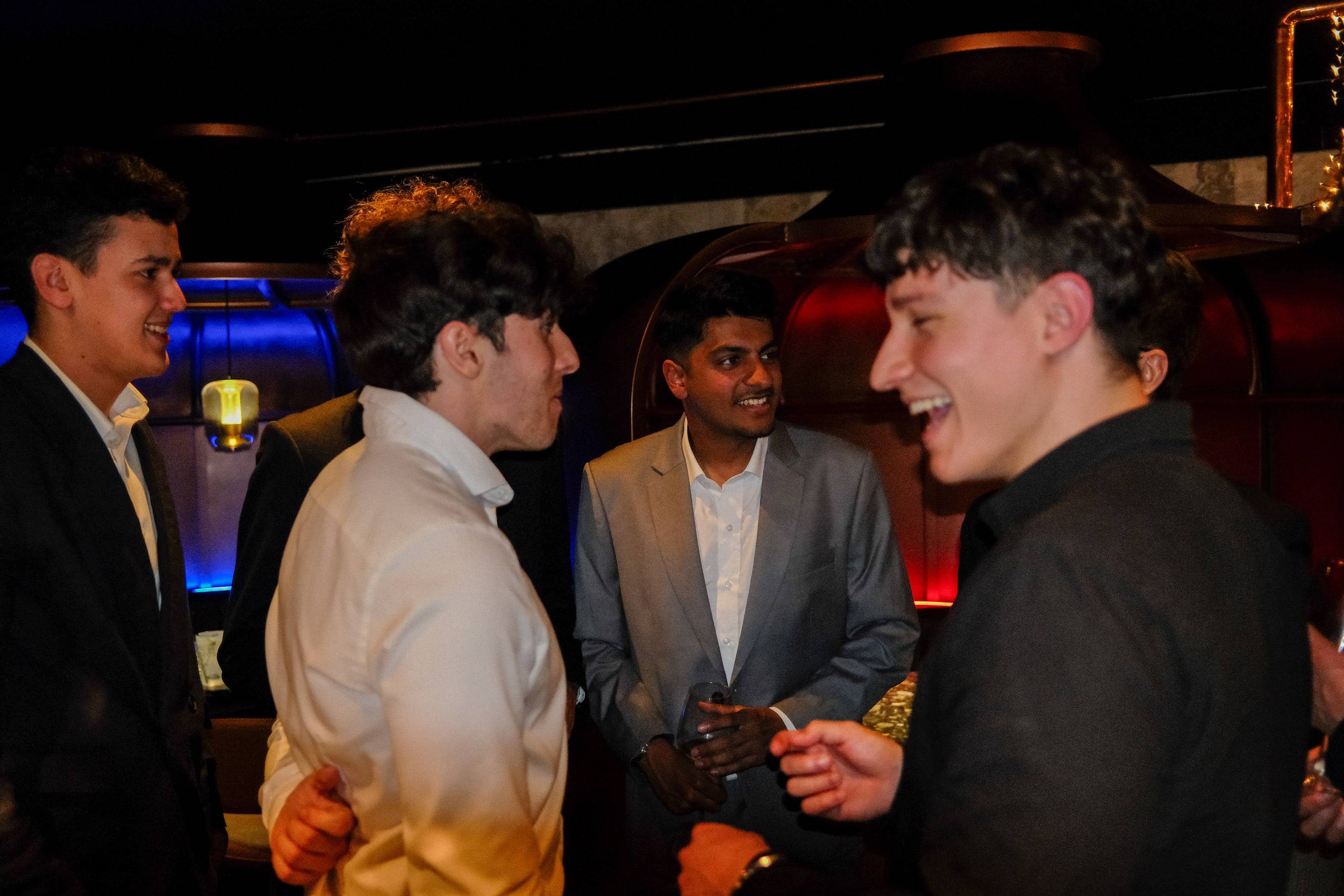 Quatre étudiants en costume discutent en riant près du bar, dans une lumière rouge et bleue.
