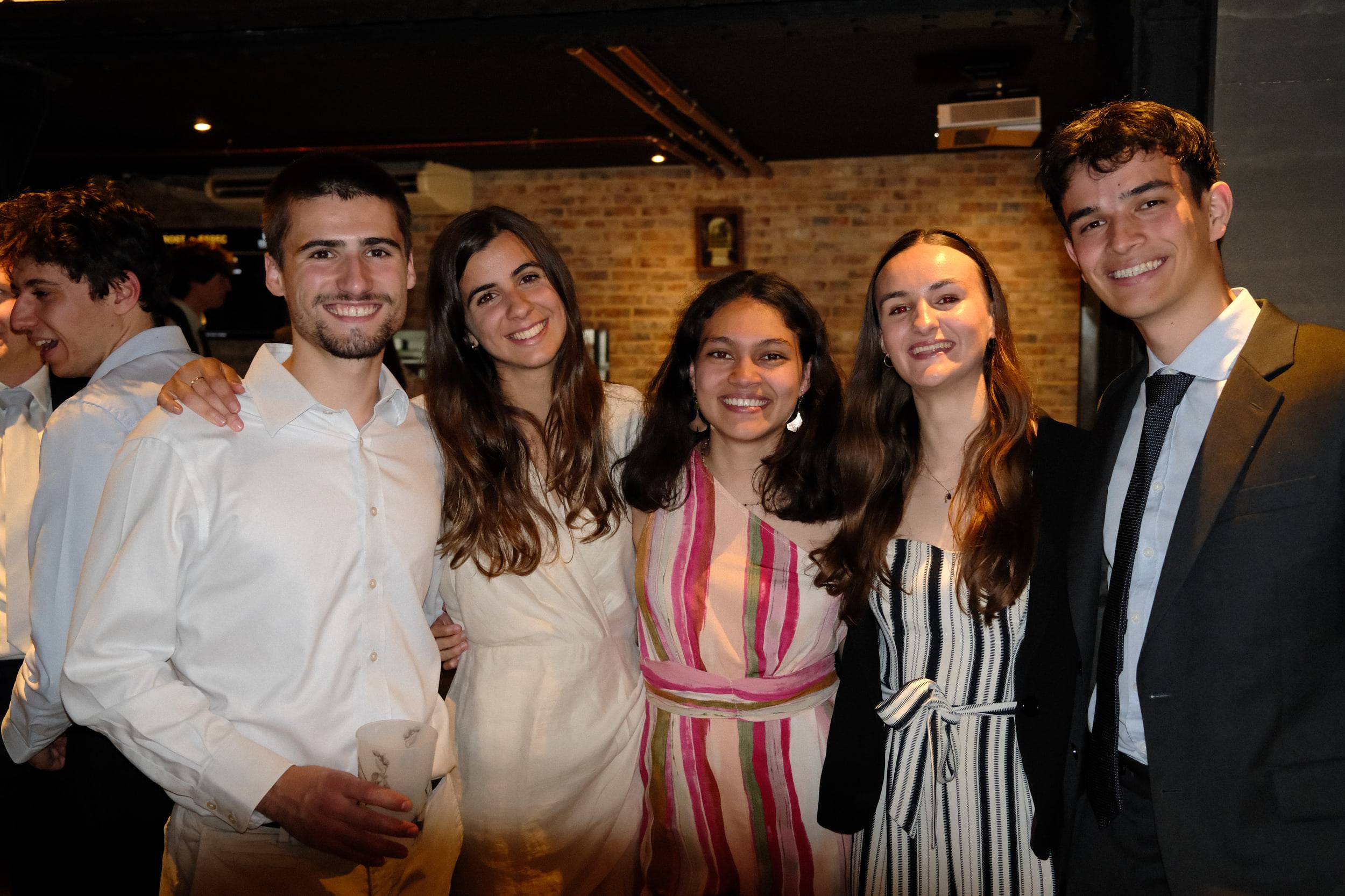 Six étudiants en tenue de soirée posent alignés devant un mur de briques éclairé.