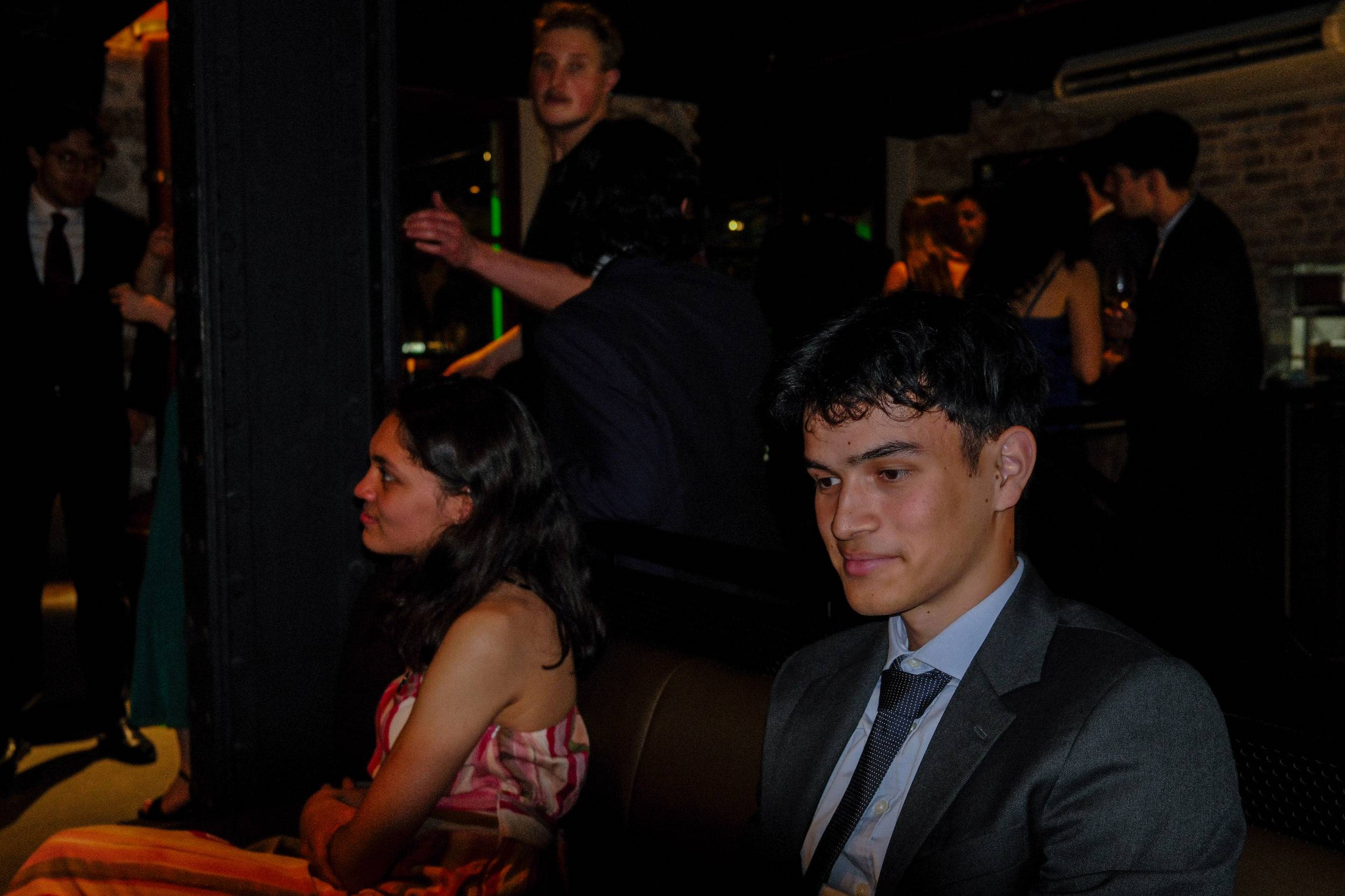 Un étudiant en costume regarde l'objectif, assis sur une banquette, une invitée de profil à côté de lui.