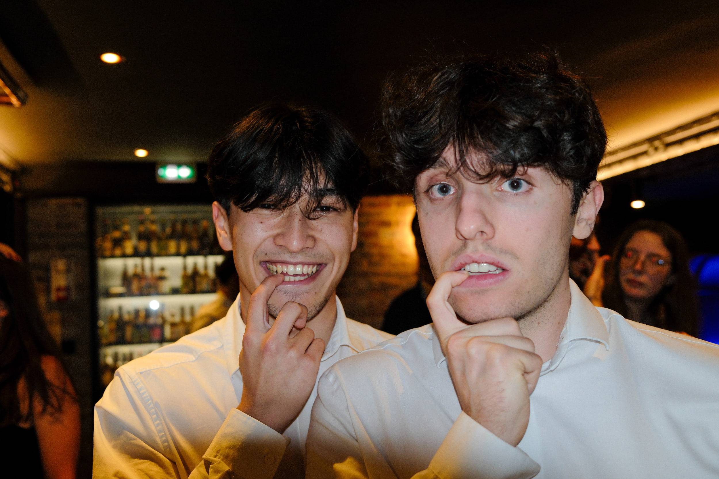 Deux étudiants en chemise blanche font une grimace face à l'objectif, le bar éclairé derrière eux.