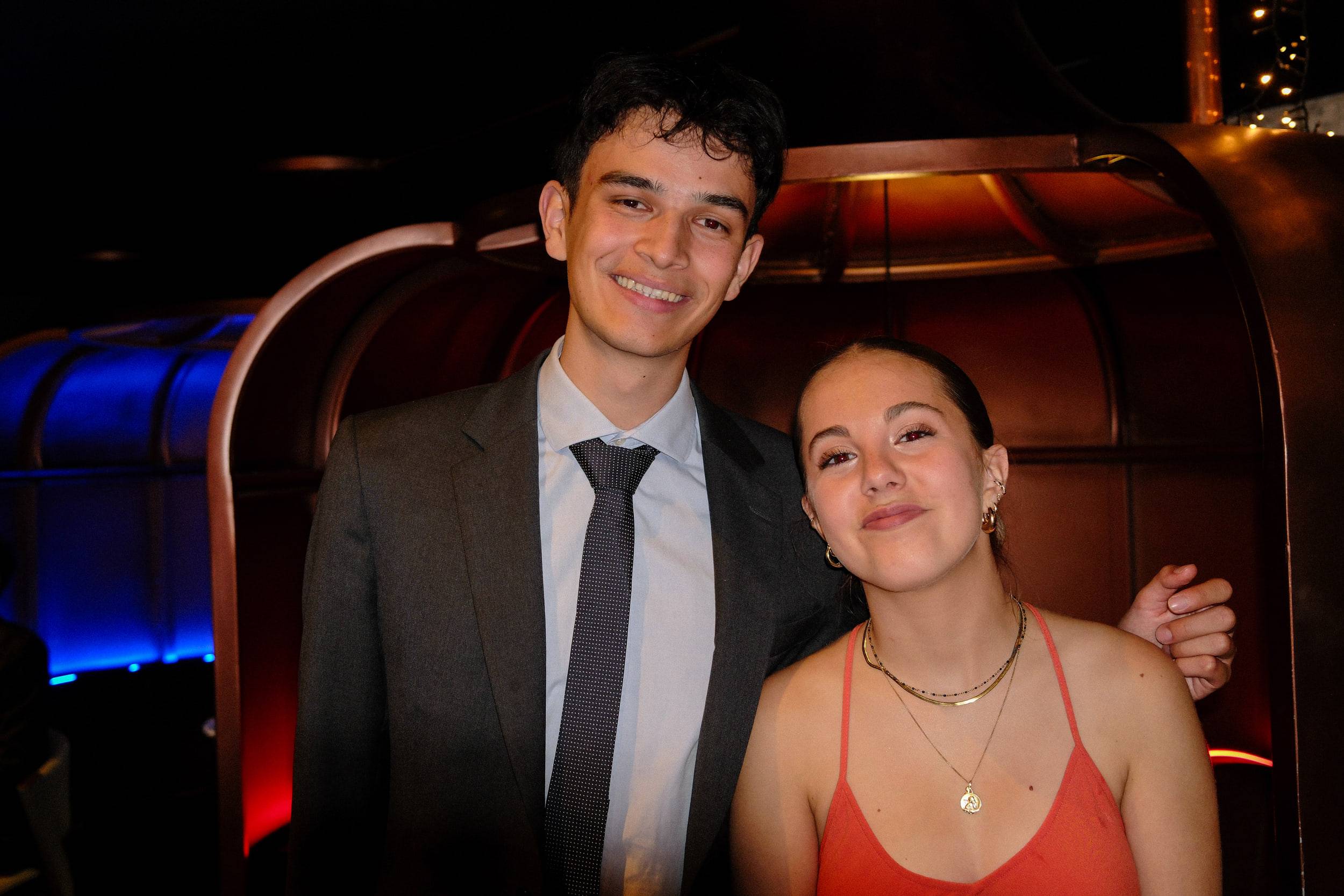 Un étudiant en costume et une étudiante en robe orange posent devant une banquette de cuir.