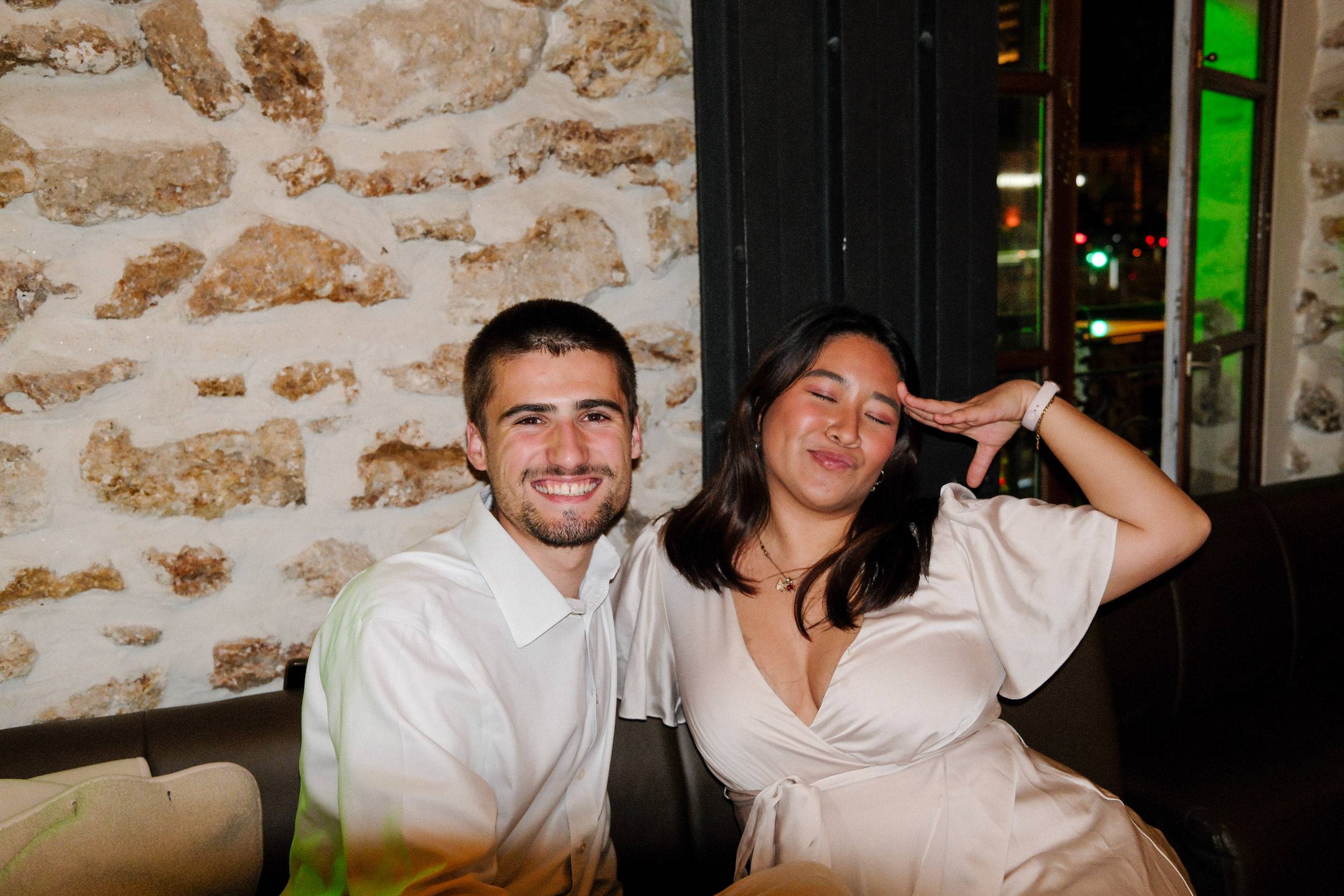Deux invités assis sur une banquette devant un mur de pierre, l'une portant la main à son front en salut.