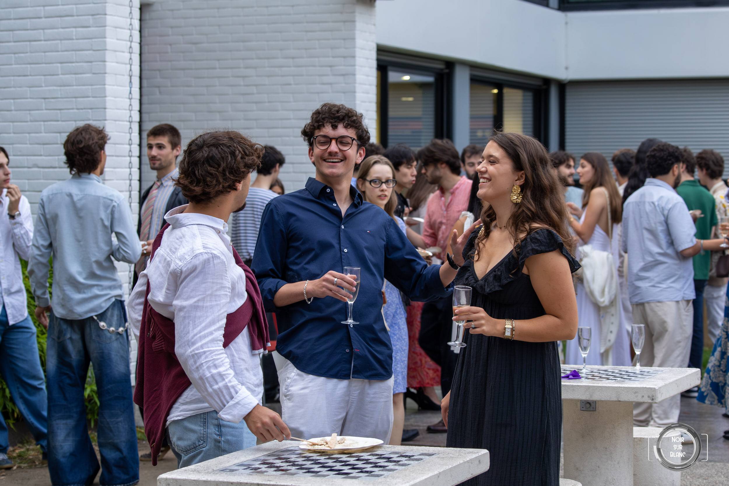 Deux invités discutent une coupe à la main autour d'un damier posé sur une table haute, pendant le cocktail en plein air.