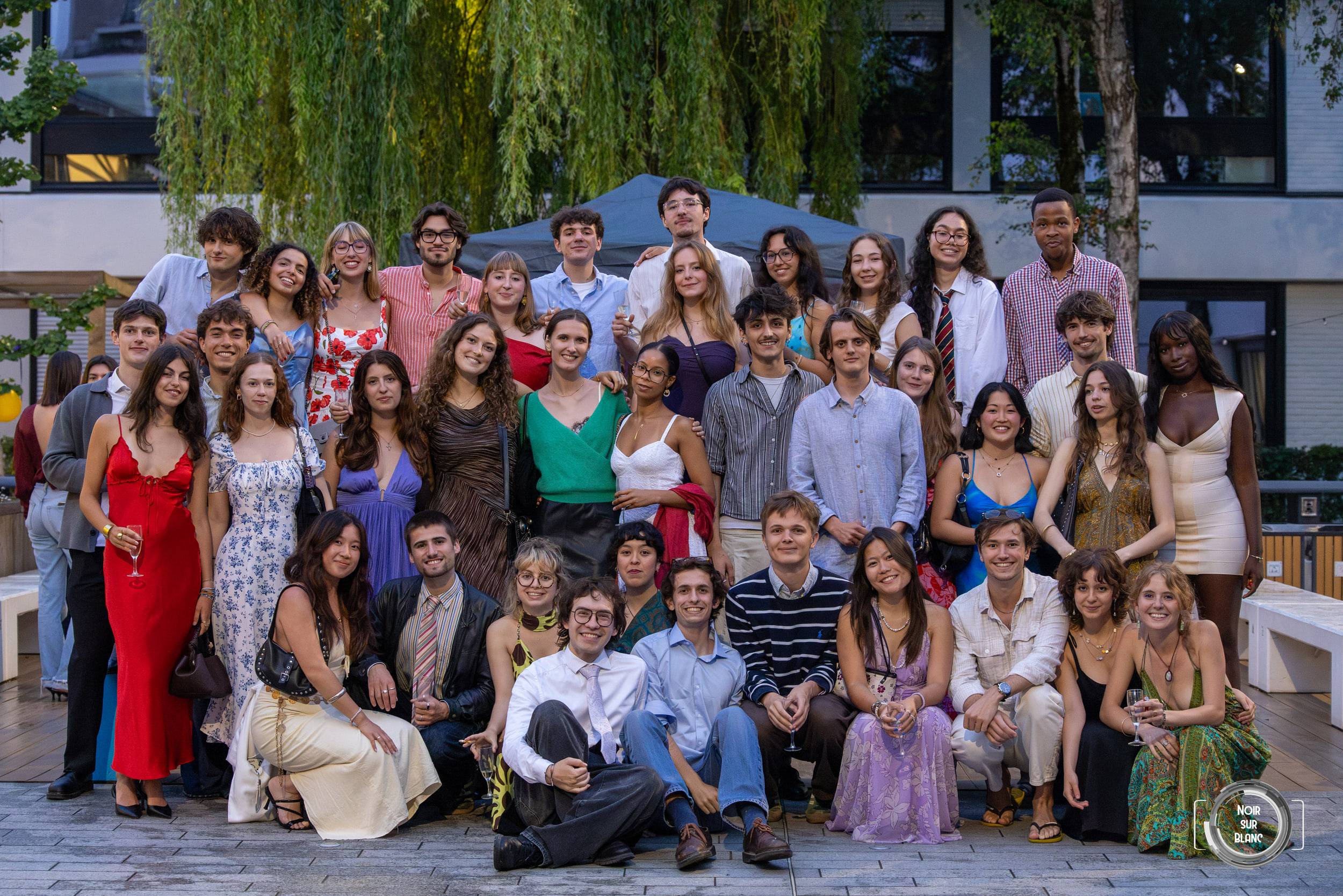 Photo de groupe d'une trentaine d'invités en tenue de soirée sur une terrasse, le premier rang assis au sol.