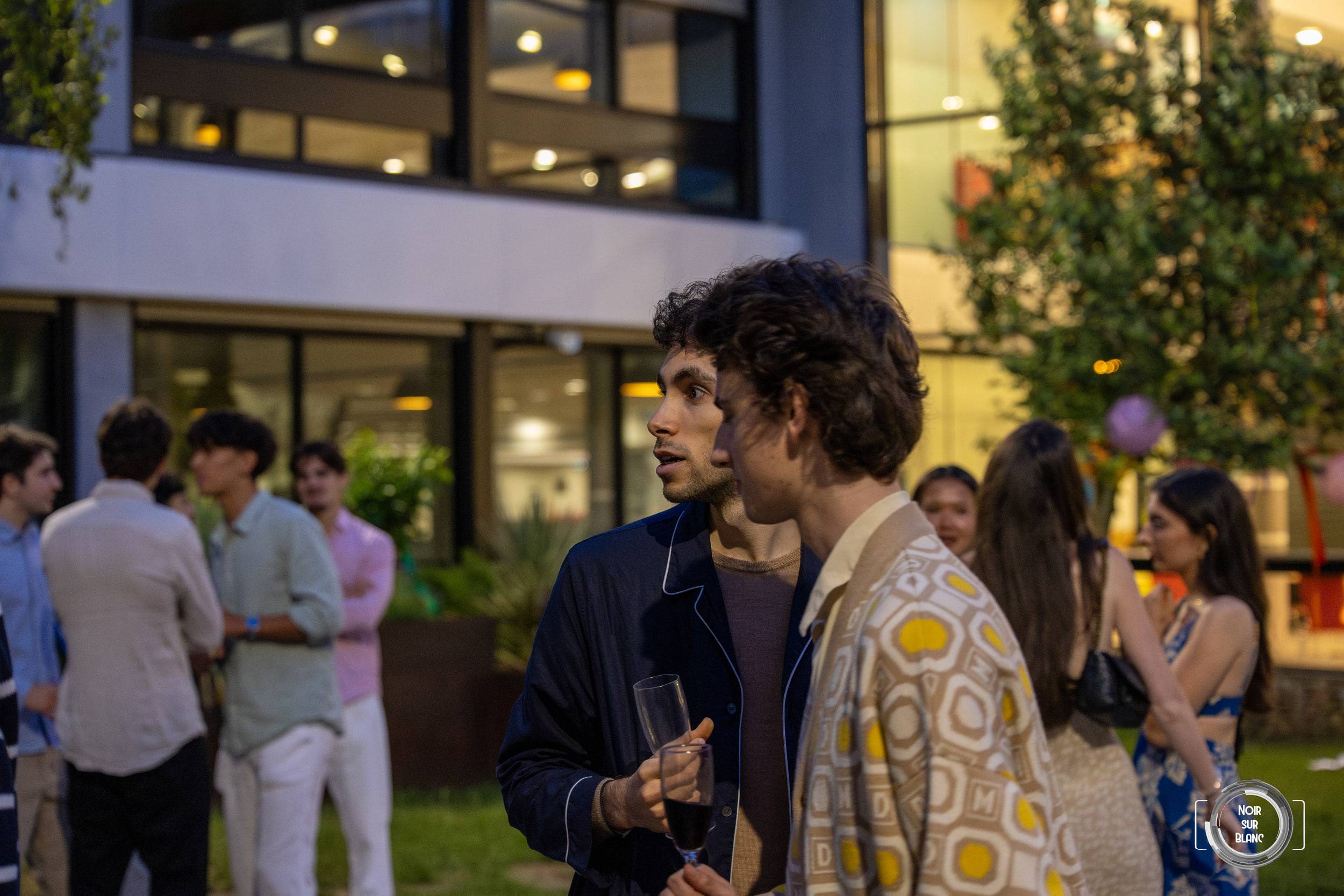 Deux invités discutent au crépuscule, l'un en veste à motifs, les convives et le bâtiment éclairé derrière eux.