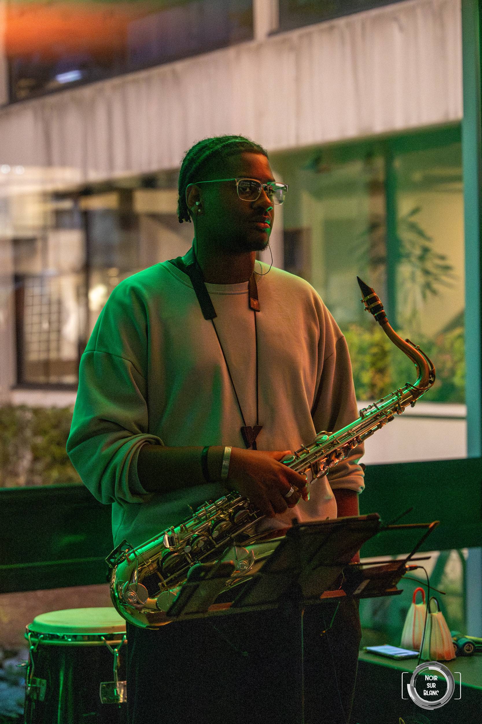 Un saxophoniste joue debout derrière ses percussions, éclairé en vert, devant la baie vitrée du bâtiment.