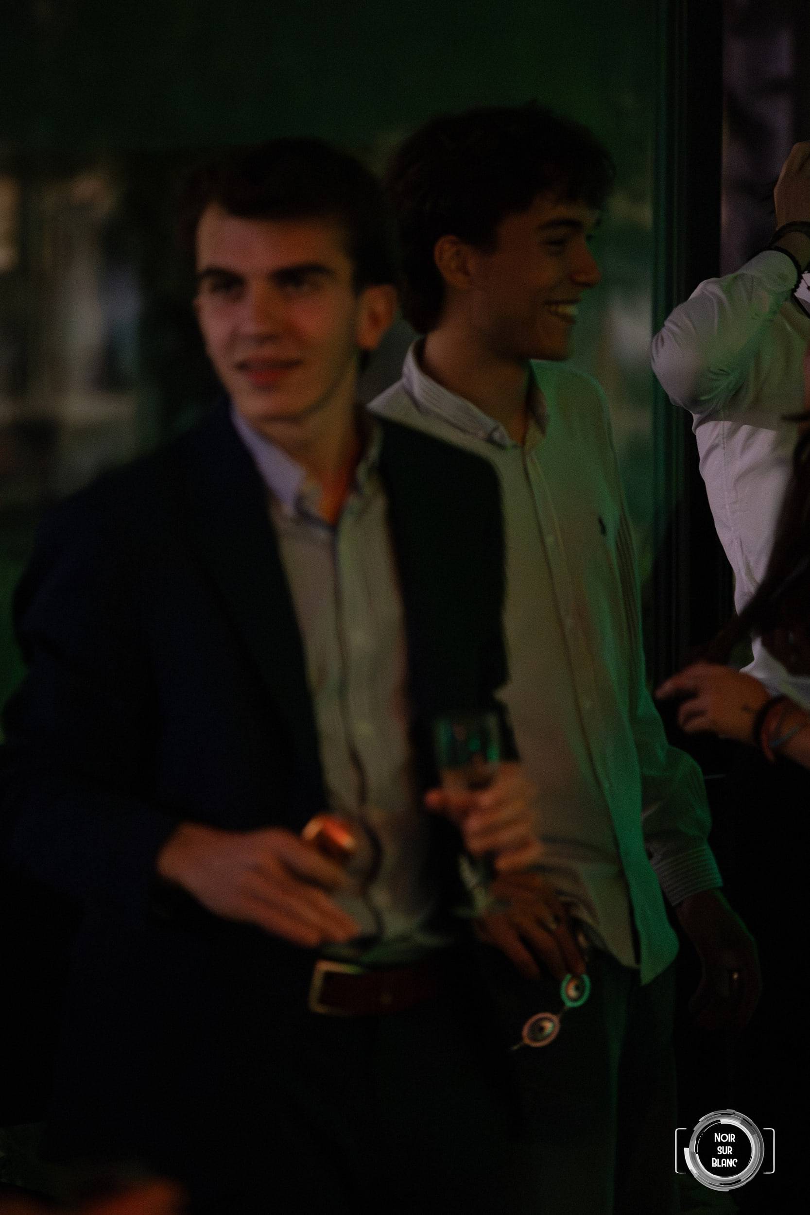 Deux invités en costume sourient côte à côte dans la pénombre, un verre à la main.