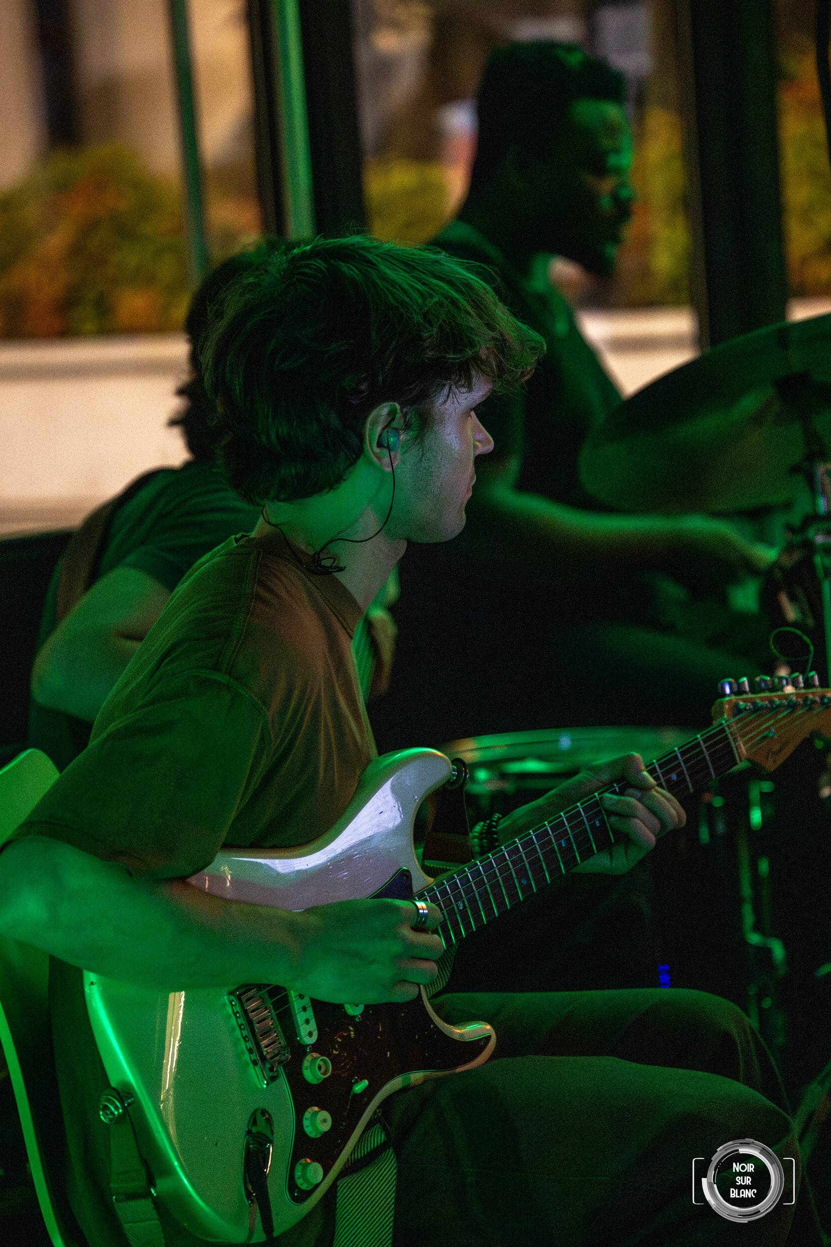 Le guitariste du groupe de jazz joue sous une lumière verte, le percussionniste derrière lui.