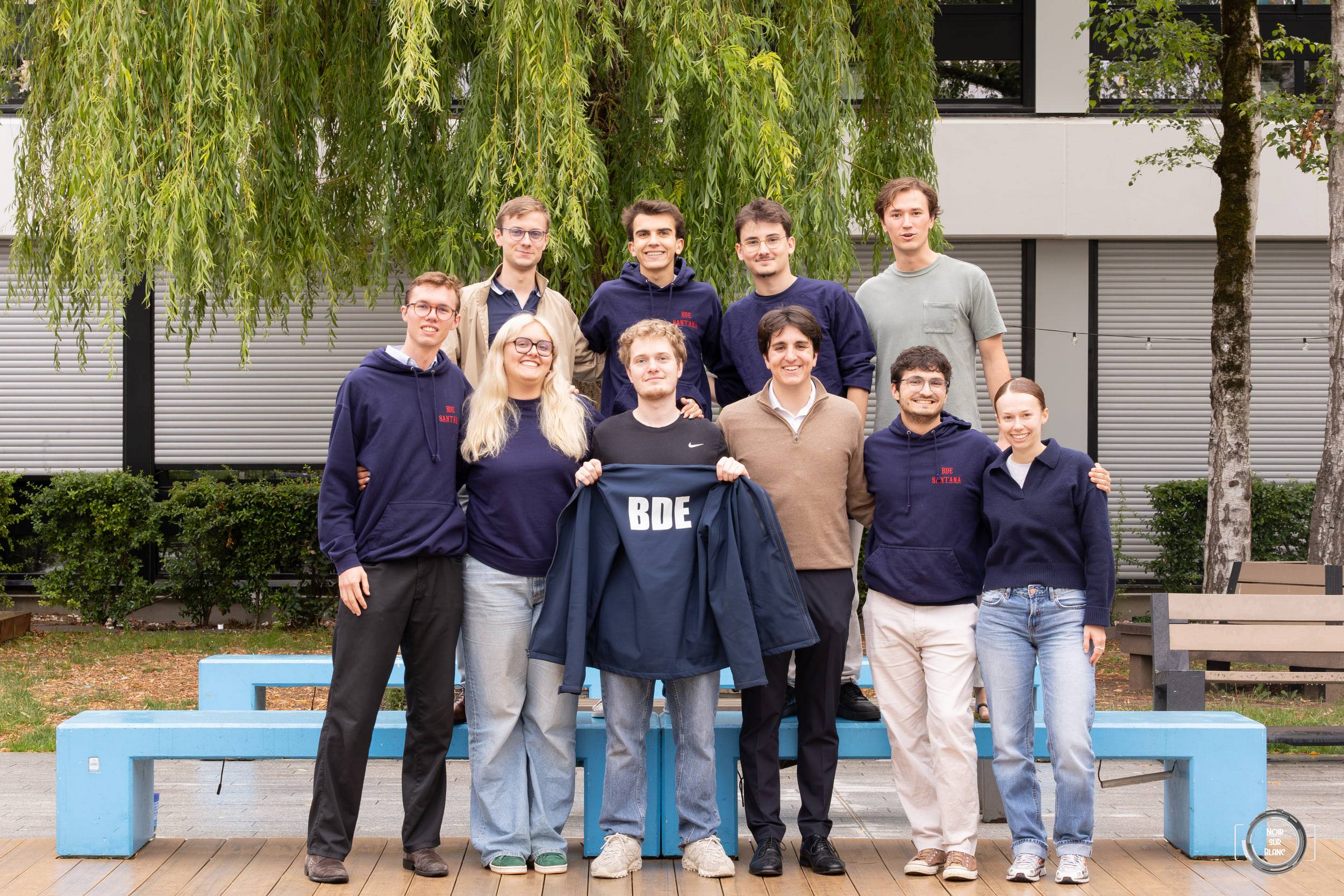 Une dizaine d'étudiants en sweat bleu marine posent en deux rangs devant des bancs bleus, deux d'entre eux tenant une veste floquée « BDE », sous un saule.