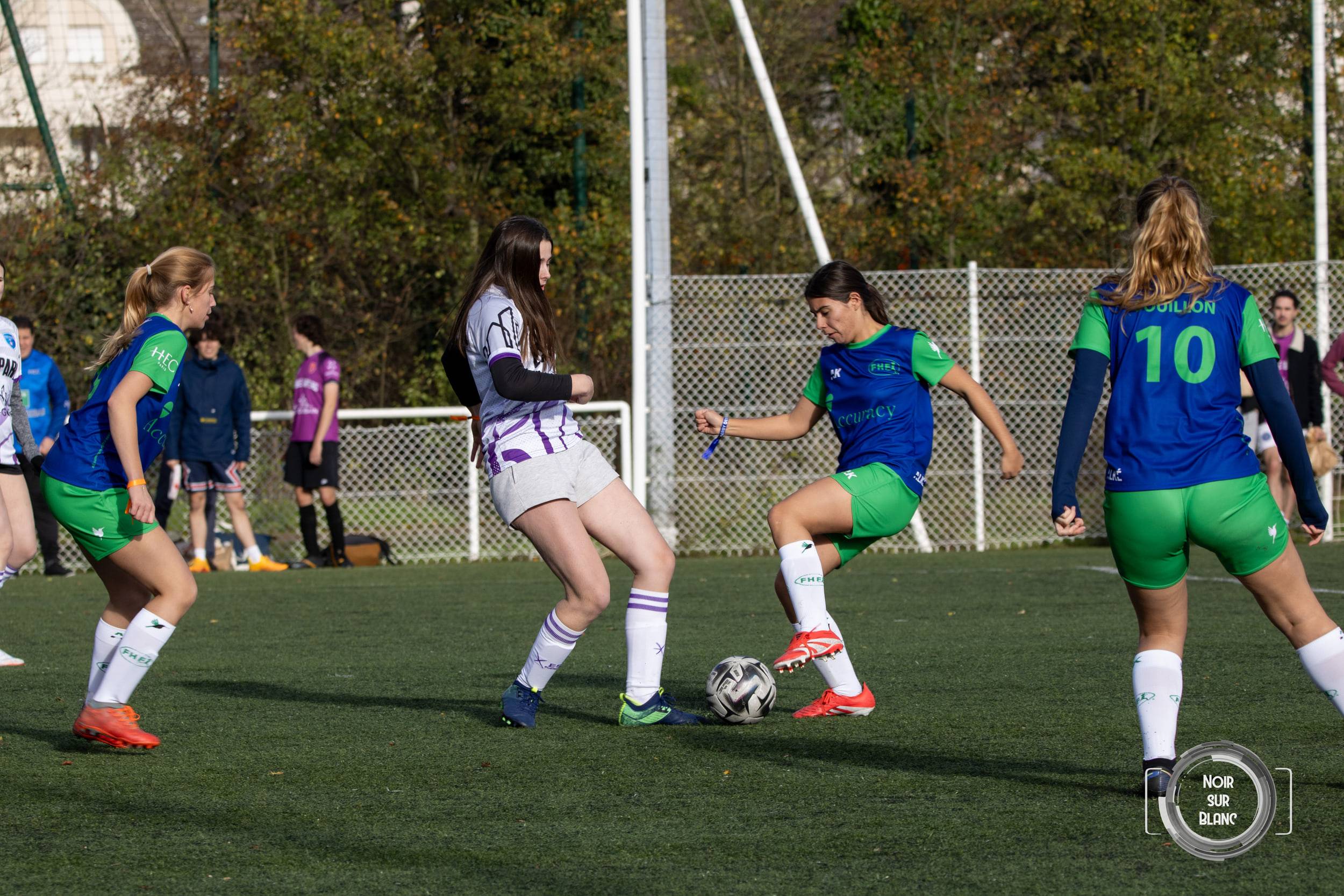 Sur la pelouse, une footballeuse en violet dispute le ballon à deux adversaires en maillot vert et bleu.